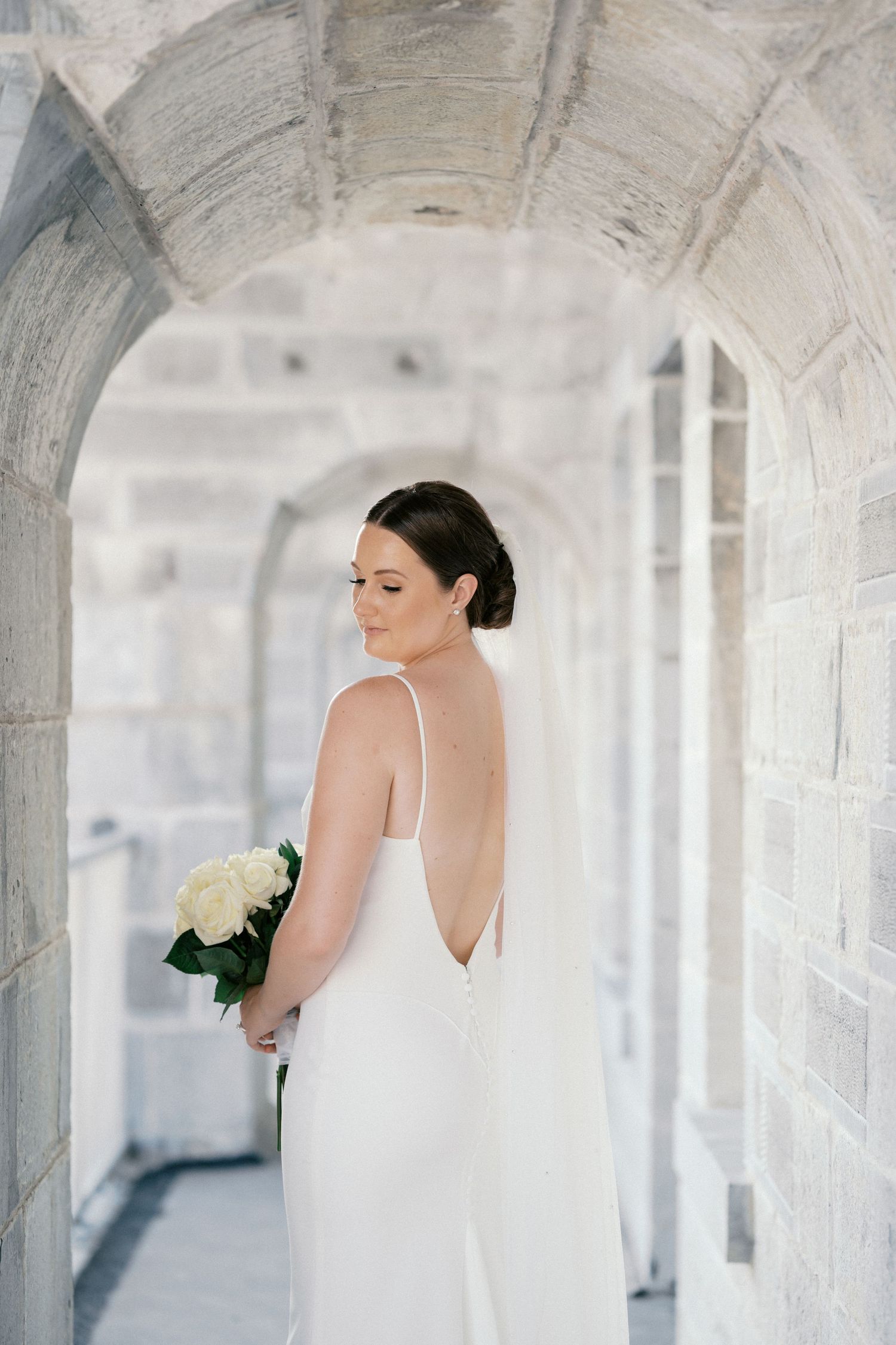 Bride in a low back white wedding gown holding white roses under stone archway.