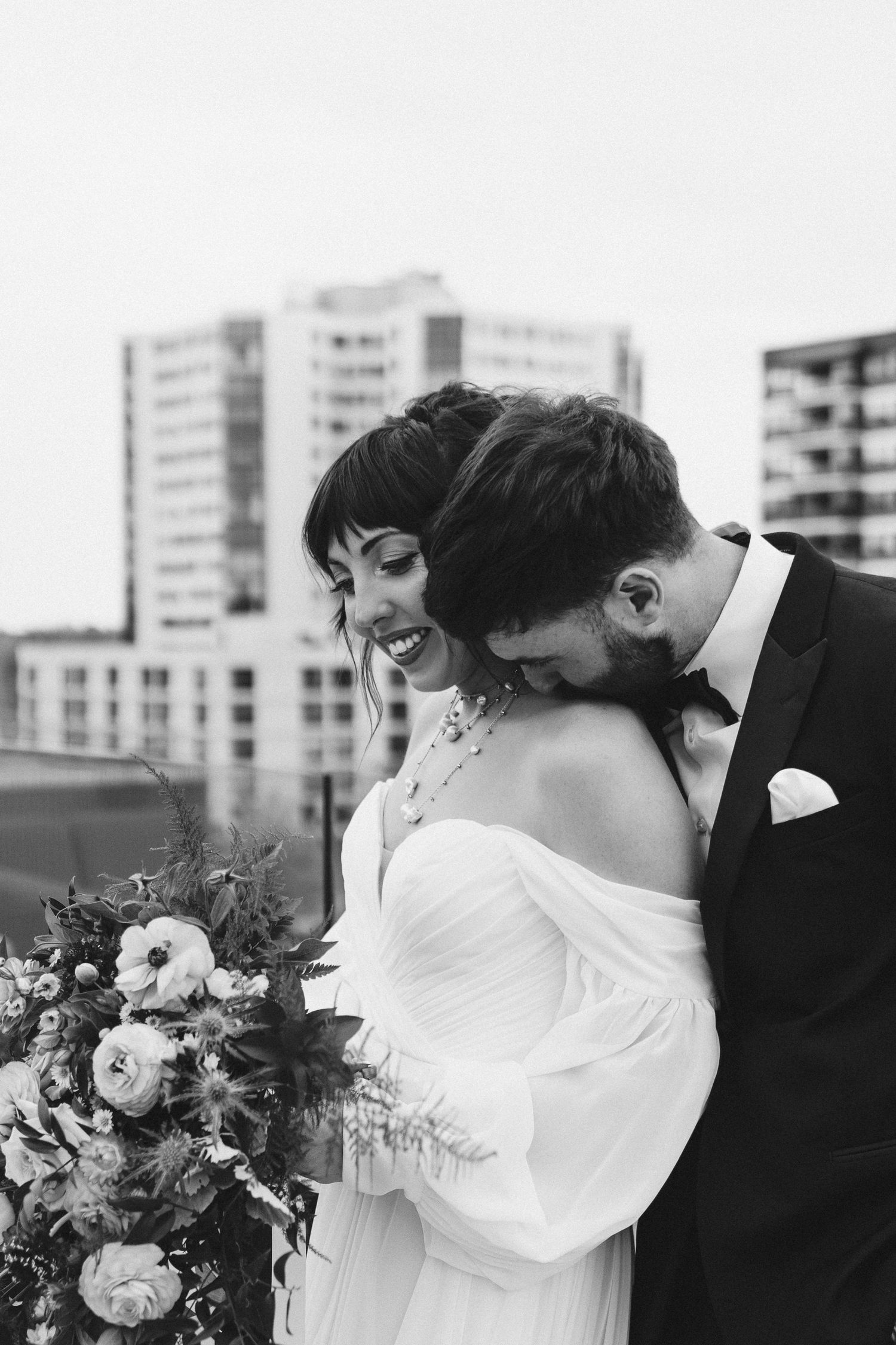 Black and white portrait of couple with cityscape backdrop and floral bouquet.