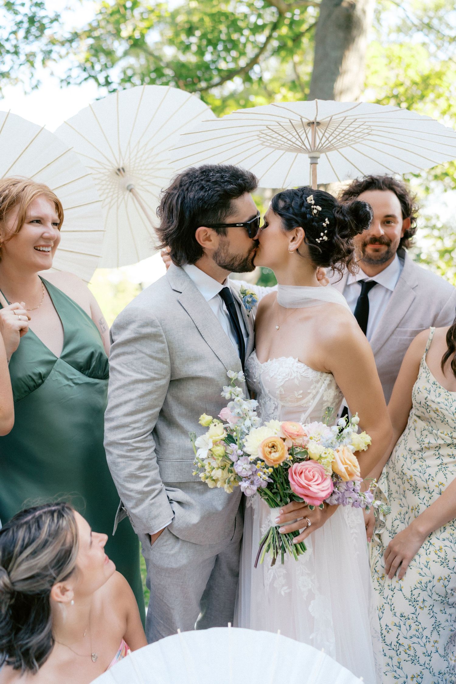 Wedding party poses with white parasols in a garden setting wearing sage green and light gray attire.
