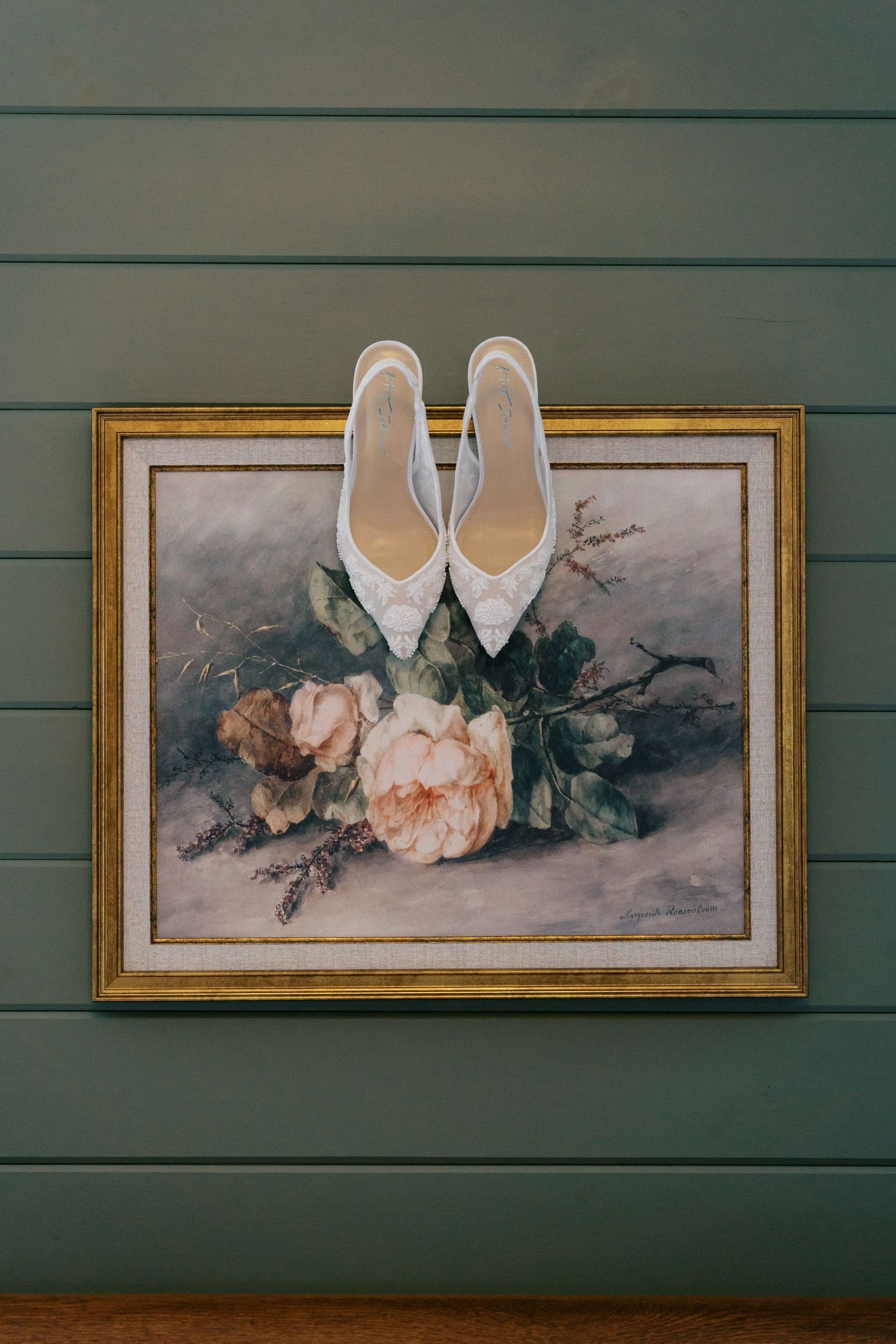 Elegant white bridal shoes displayed against sage green wall with vintage floral artwork in gold frames.