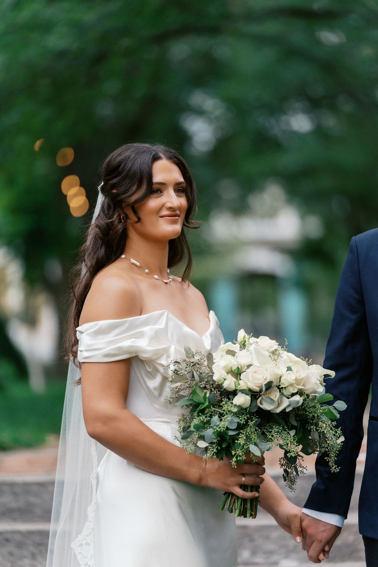 A bride in an off-shoulder white gown holds a lush bouquet of white flowers during an outdoor wedding.