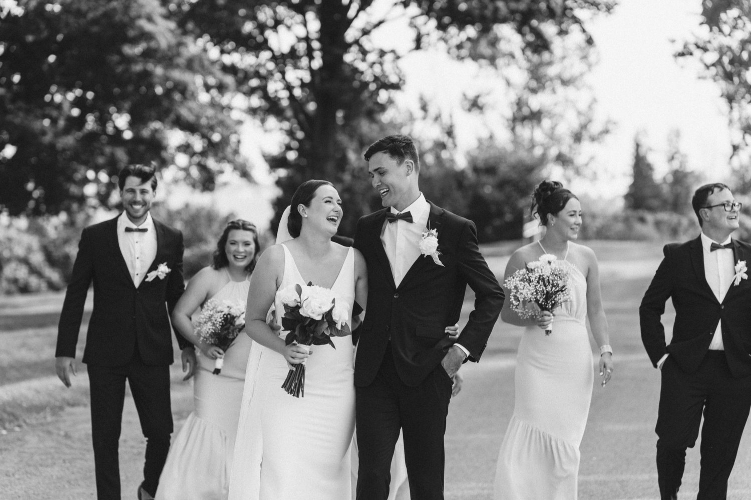 Black and white photo of a wedding party walking outdoors, bride and groom laughing together with bridesmaids and groomsmen.