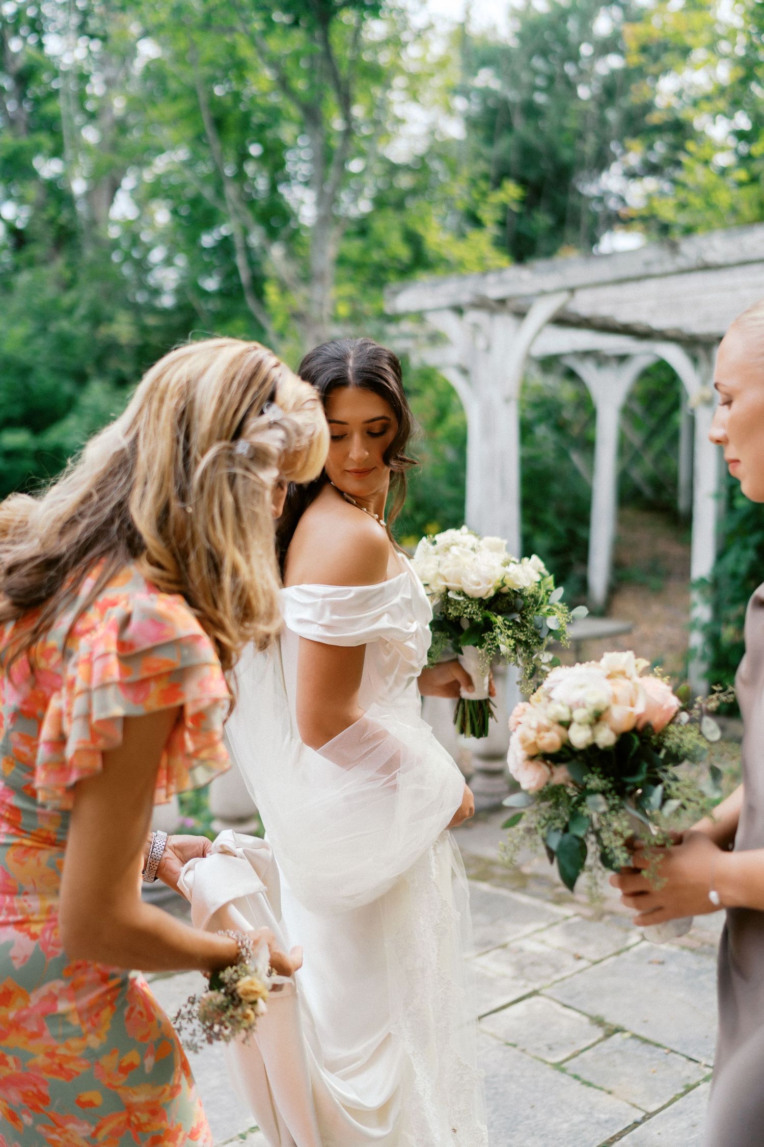 Wedding party members gather around a bride in an elegant white dress holding bouquets in a garden setting.