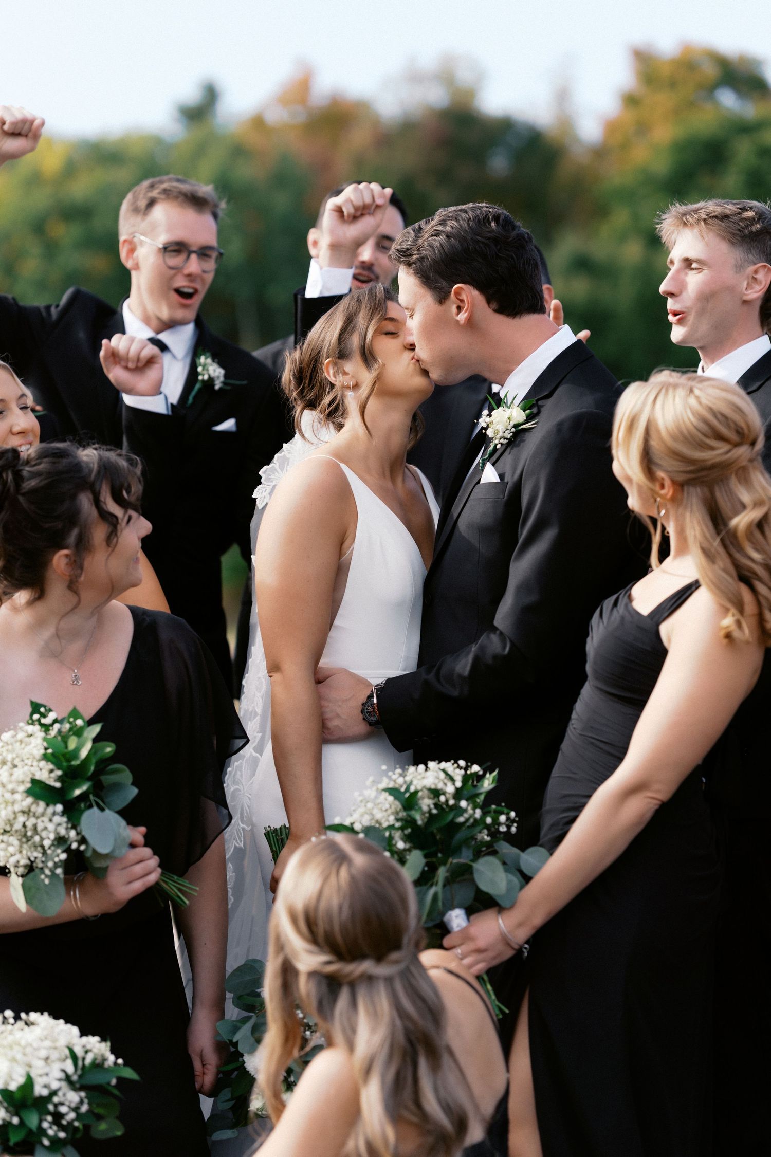 Wedding party laughs together outdoors wearing formal black attire and holding bouquets.