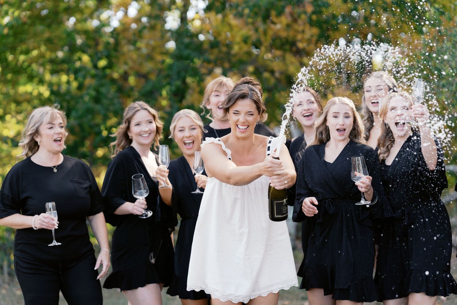 Wedding party in black dresses celebrating outdoors with champagne and laughter.