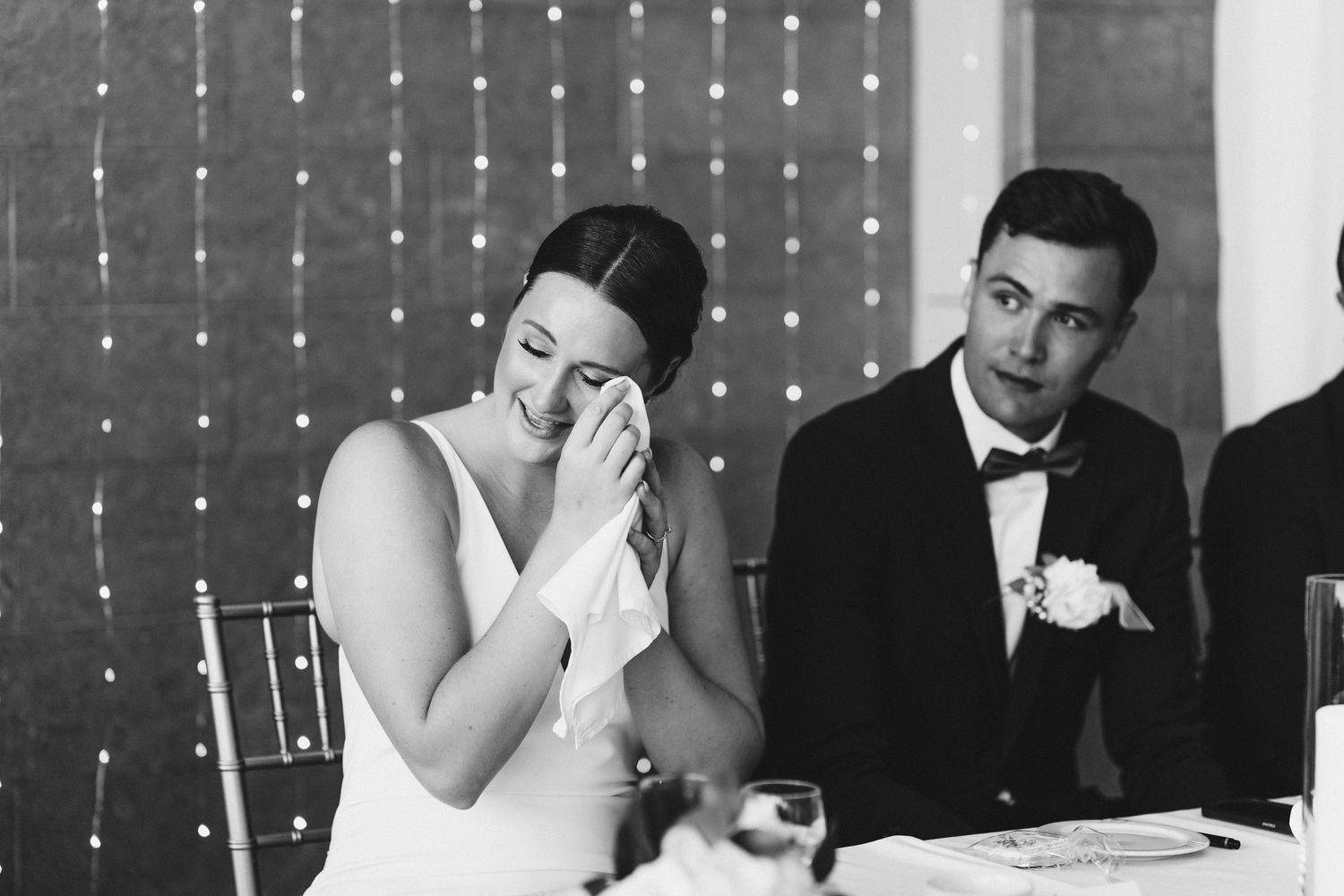 Emotional bride wiping tears beside groom in tuxedo during wedding reception speech.