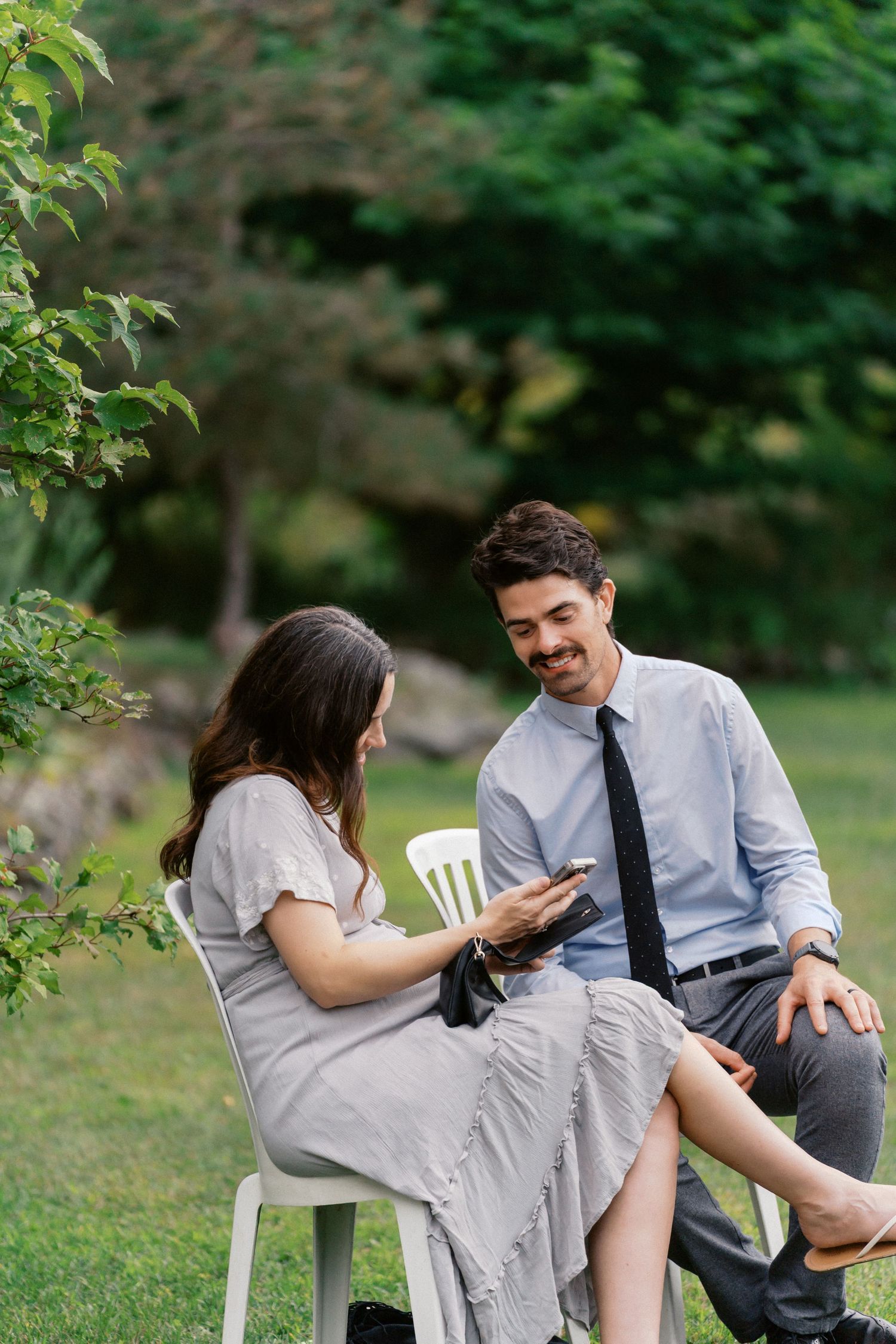 A couple sits together on white chairs in a garden setting looking at their phones.