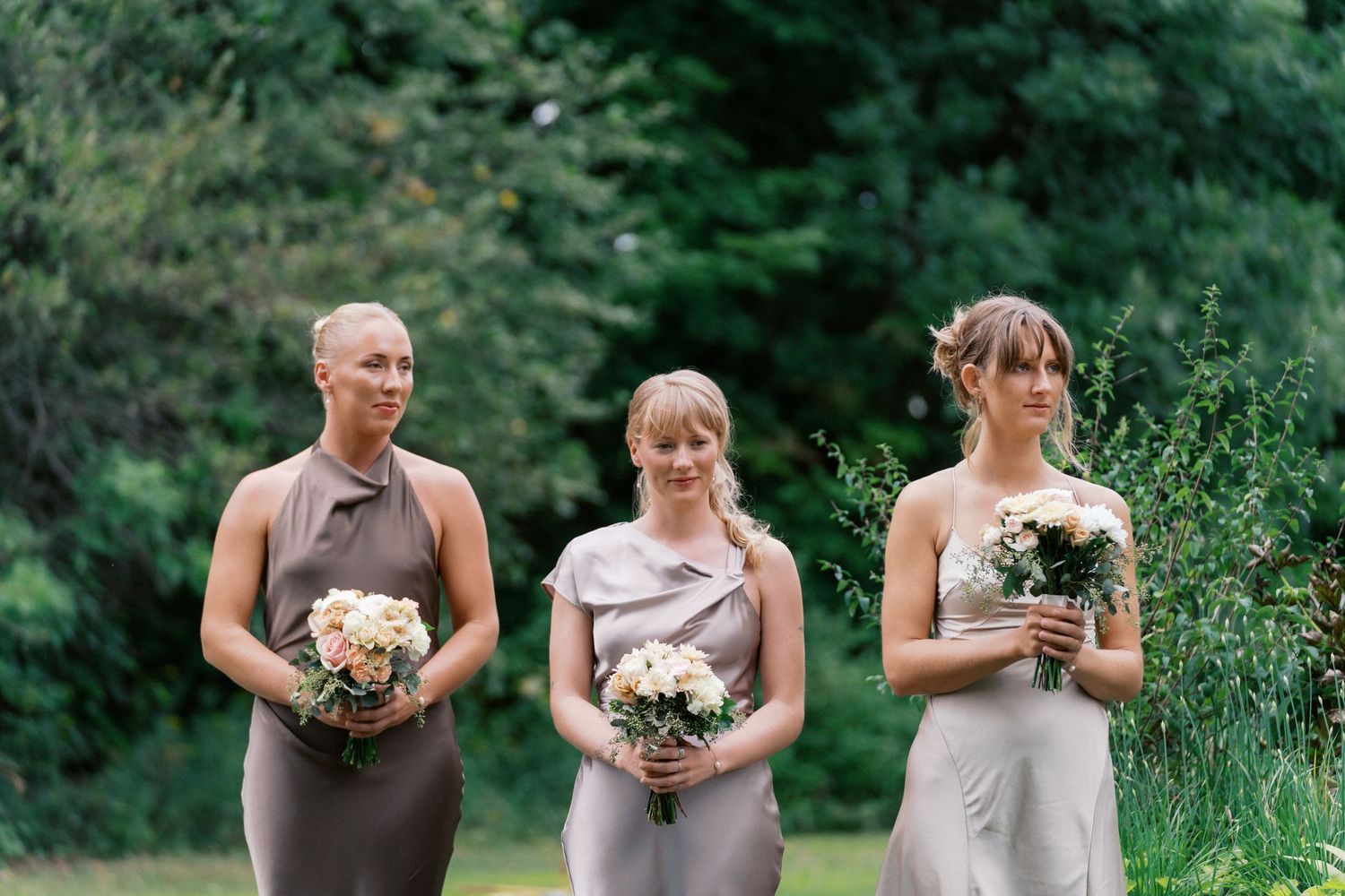 A row of bridesmaids in gray dresses holding white floral bouquets stand together in a garden setting.