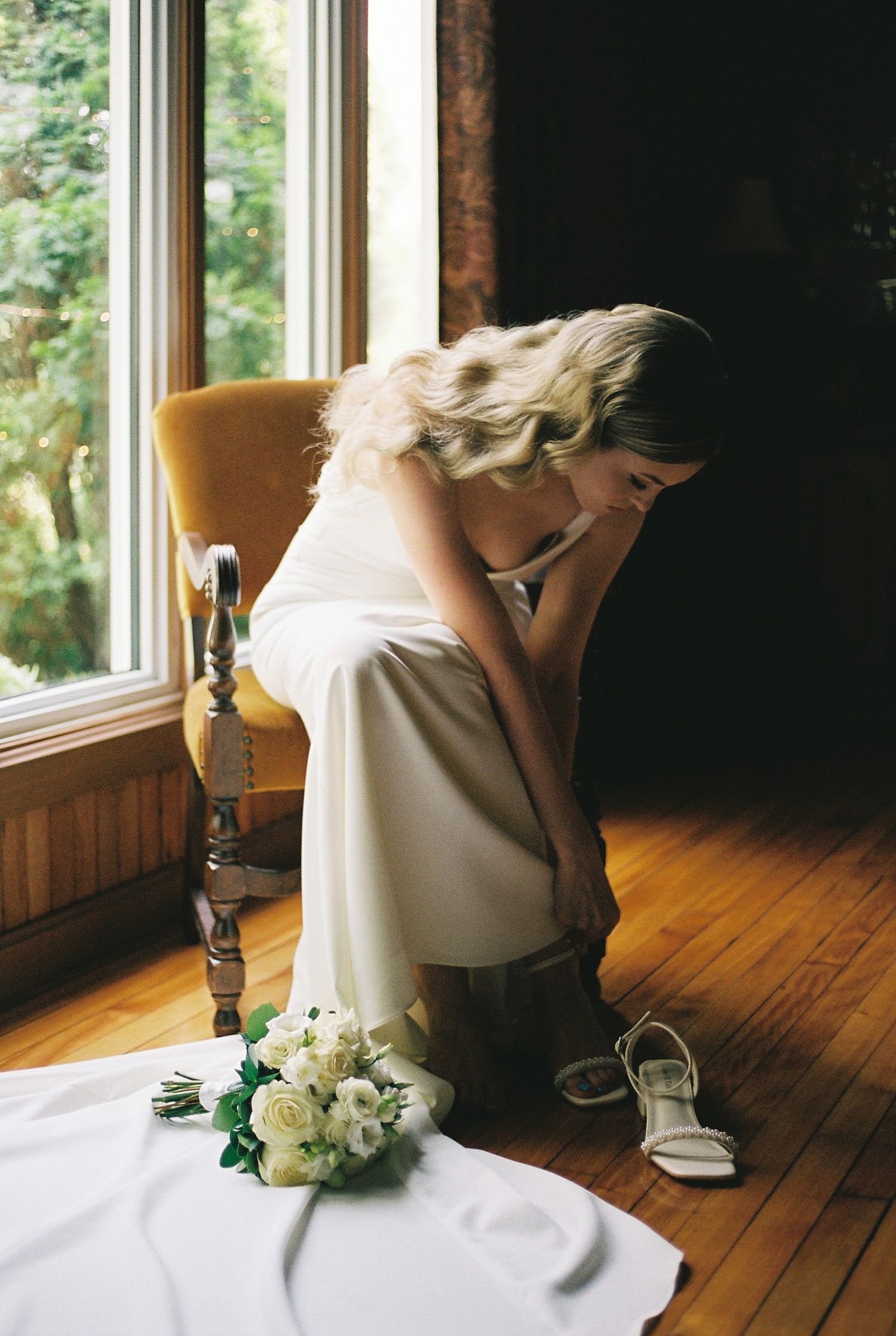 Bride getting ready near a window with white flowers and shoes on the floor.