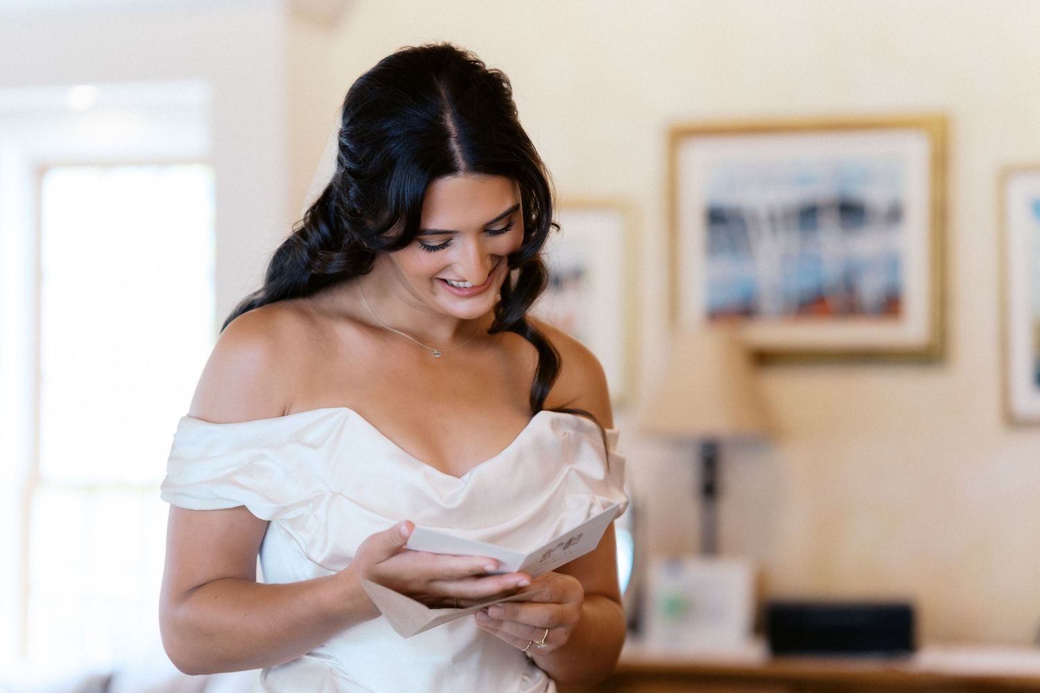 A person in a white off-shoulder dress smiles while looking down at something in their hands.