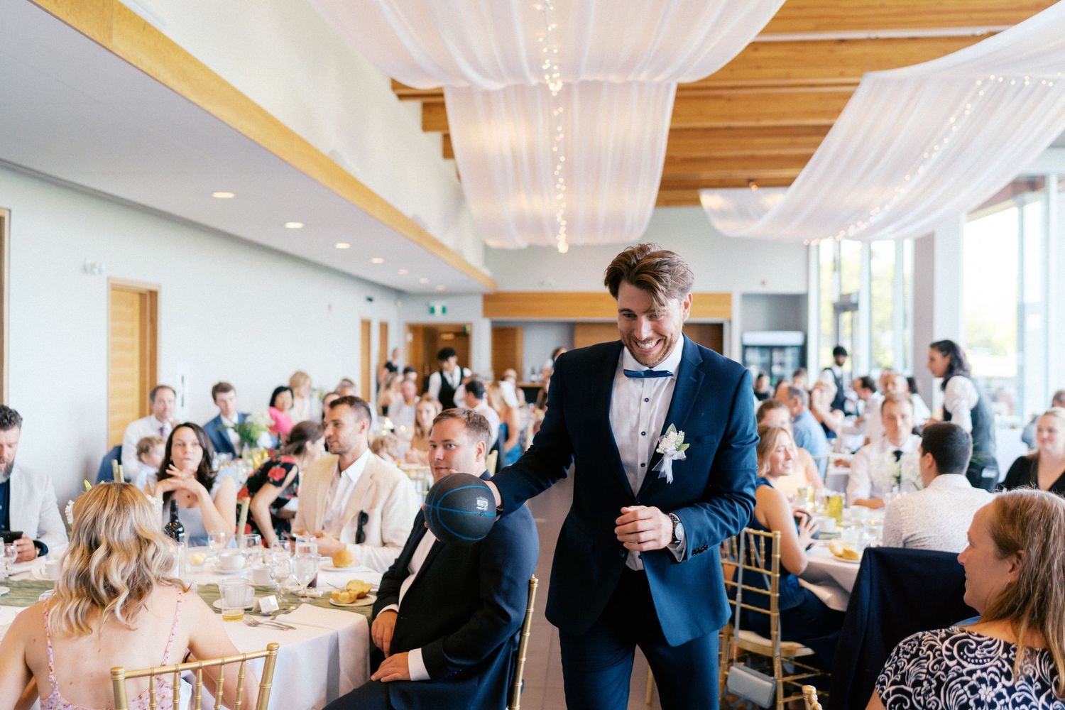 Groom in navy suit giving a speech at elegant wedding reception with draped ceiling and seated guests.
