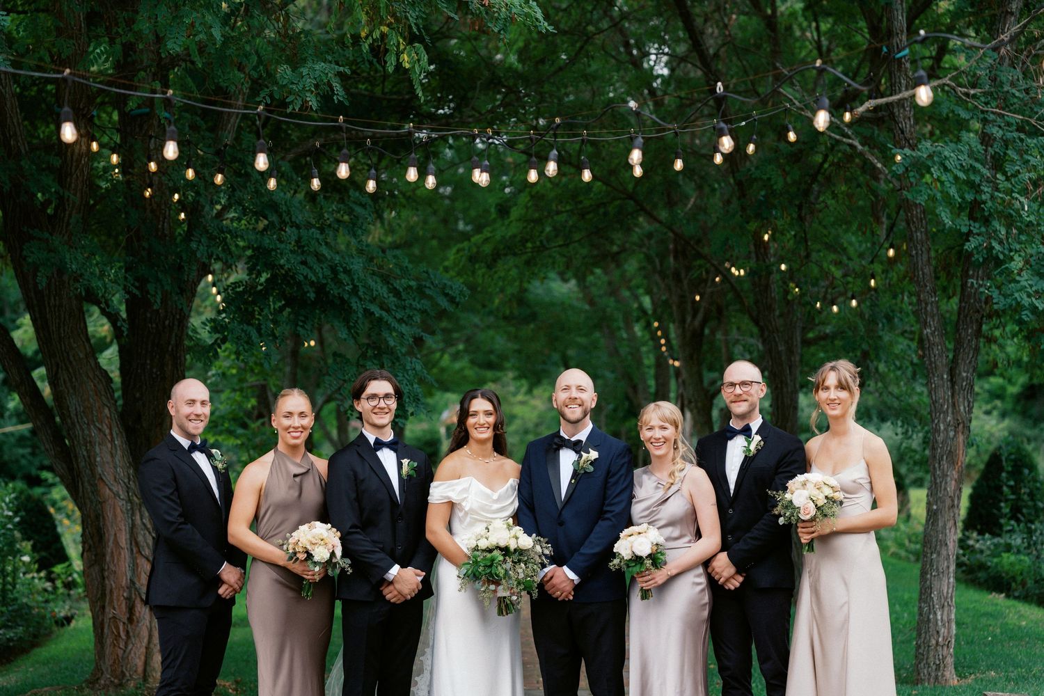 Wedding party poses under string lights and trees in formal attire.