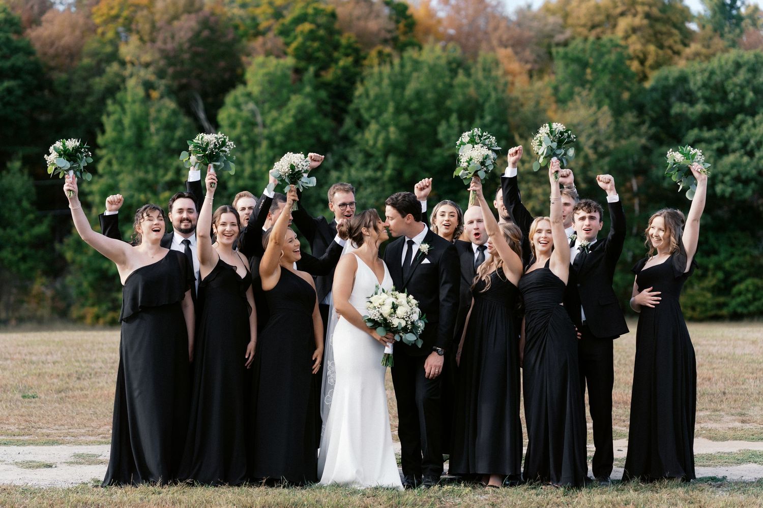 Wedding party celebrating outdoors with joyful expressions against natural backdrop.