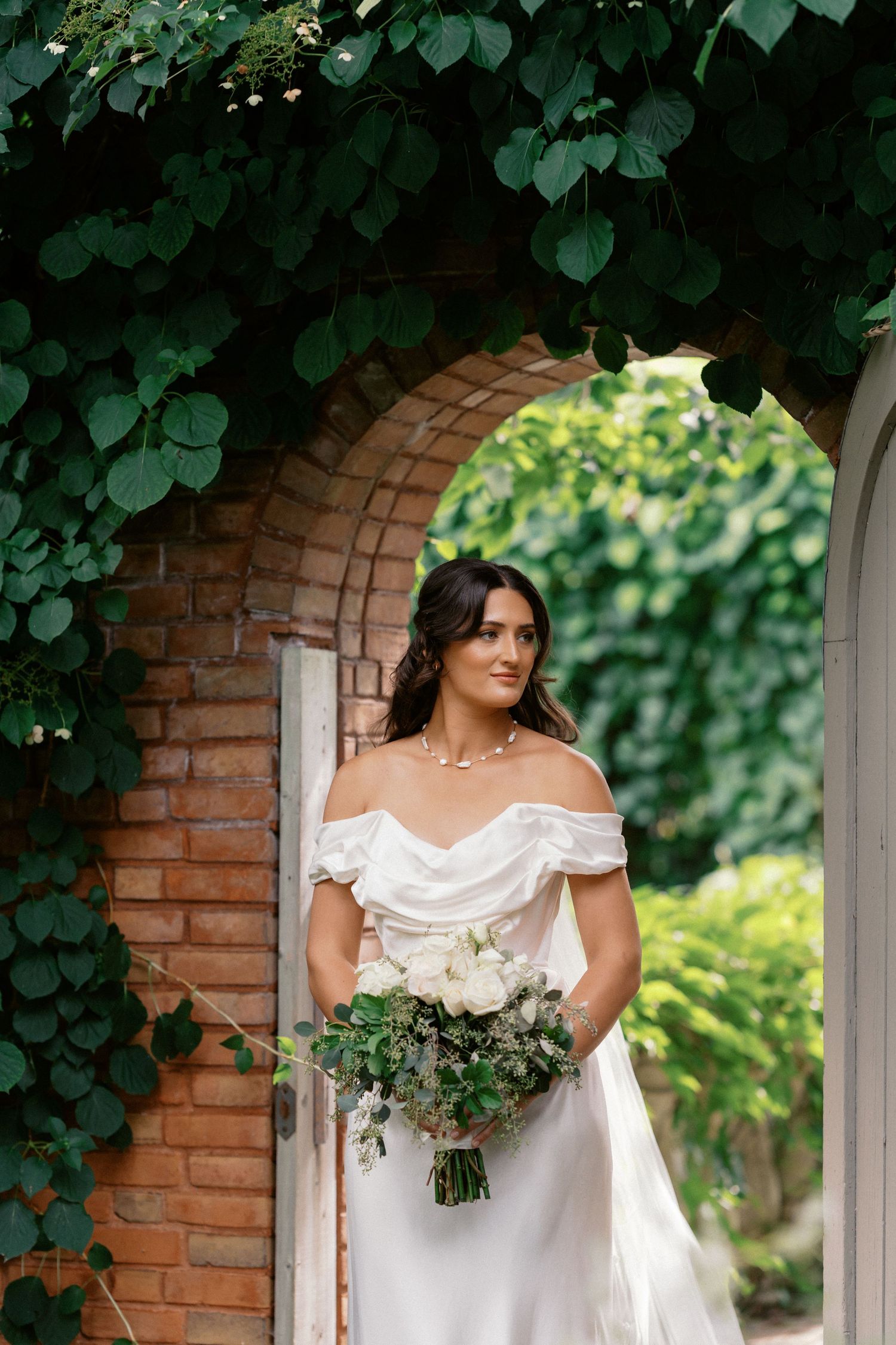 White wedding dress with off-shoulder detail framed by brick archway and green foliage.
