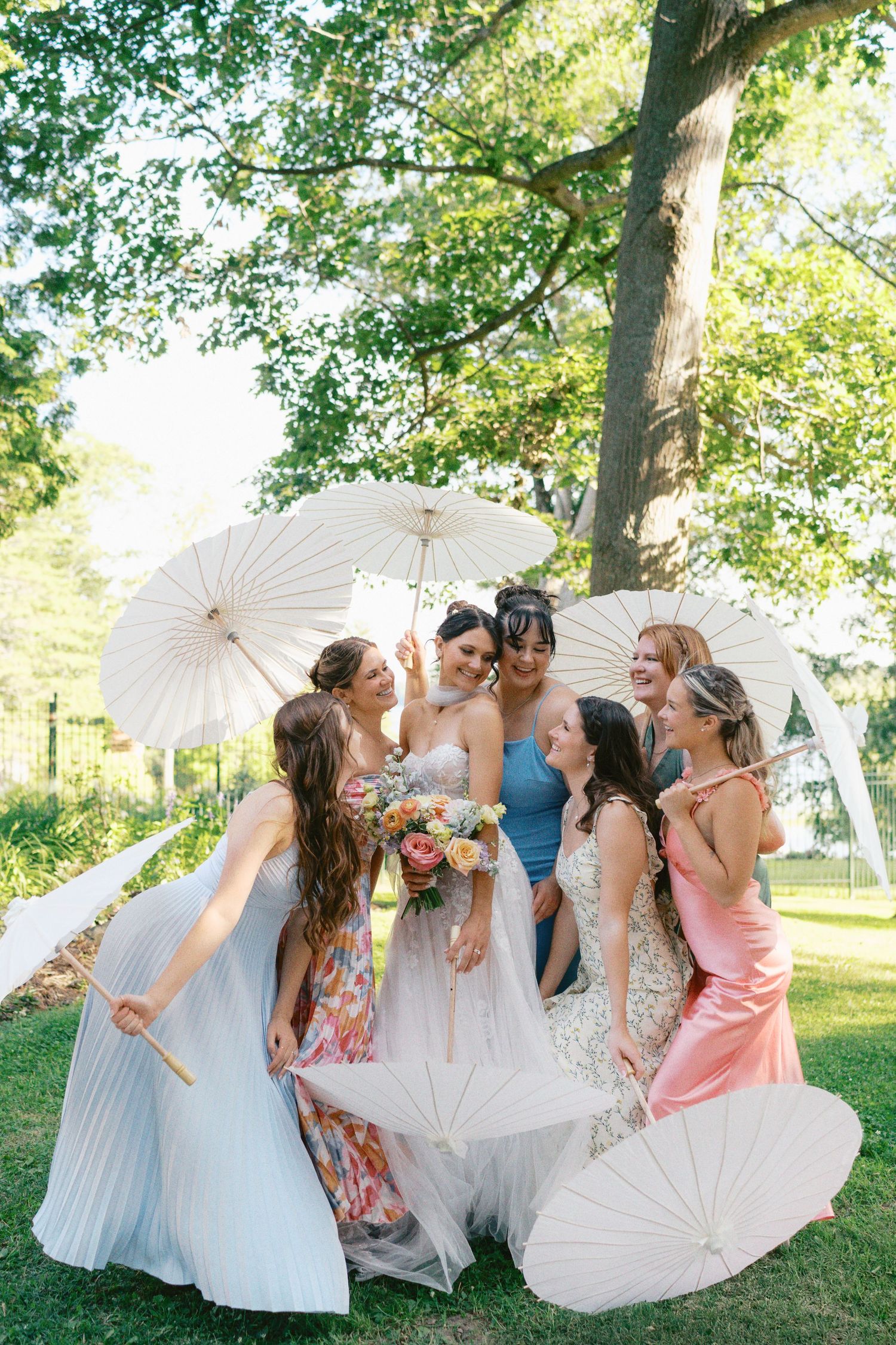 A group of wedding party members pose playfully with white parasols in a sunny outdoor garden setting.