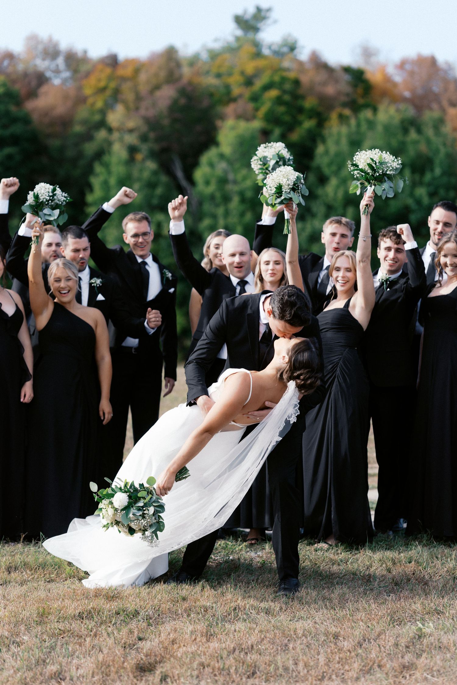 Wedding party celebrating with raised arms against autumn foliage backdrop.