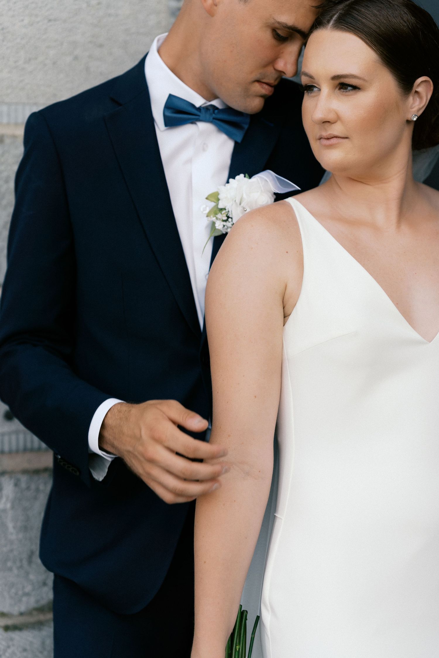 Bride and groom in elegant wedding attire, groom in navy suit with teal bow tie embracing bride in white gown.