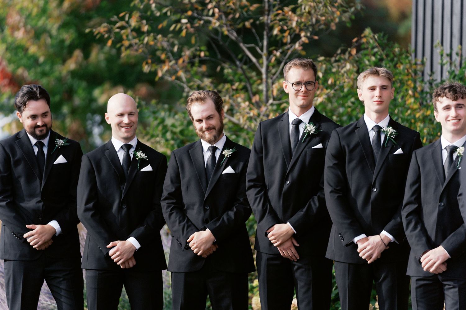 Groomsmen in black suits stand in a row outdoors during a formal wedding ceremony.