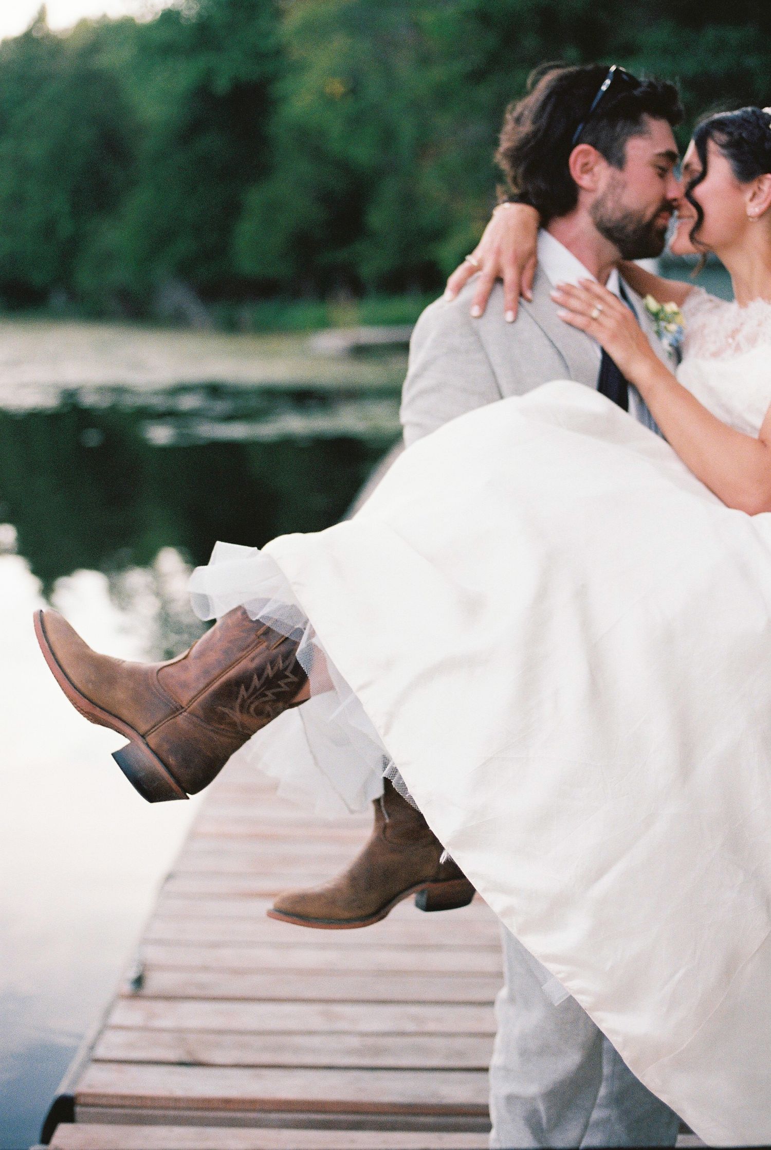 A romantic wedding couple sits on a wooden dock by the water, the bride's cowboy boots visible in a candid moment.
