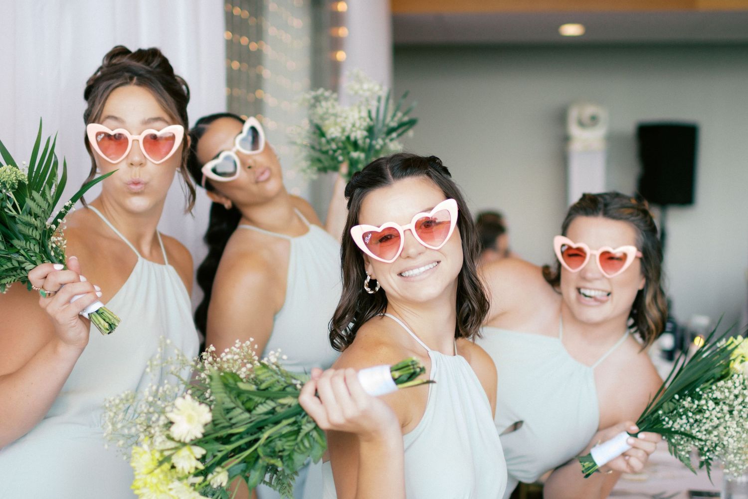 Bridesmaids in white dresses wearing heart-shaped sunglasses and holding floral bouquets at a wedding.