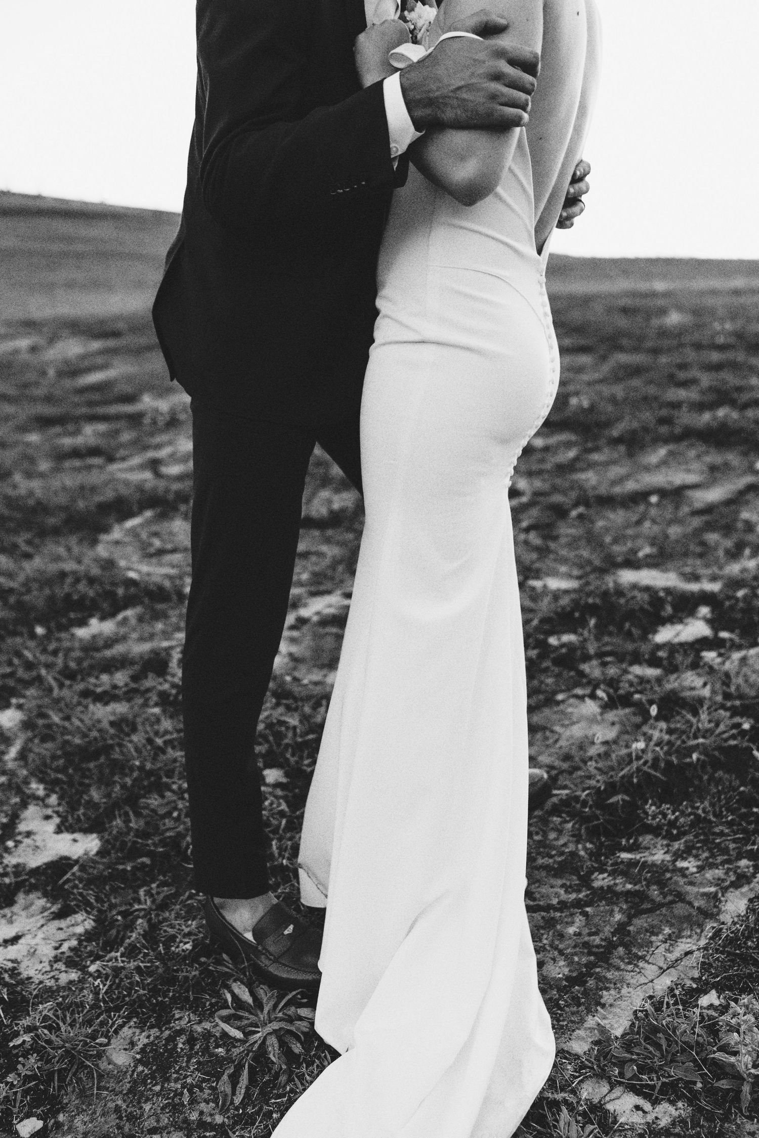 Black and white photo of bride in white gown and groom in dark suit embracing outdoors in a field.