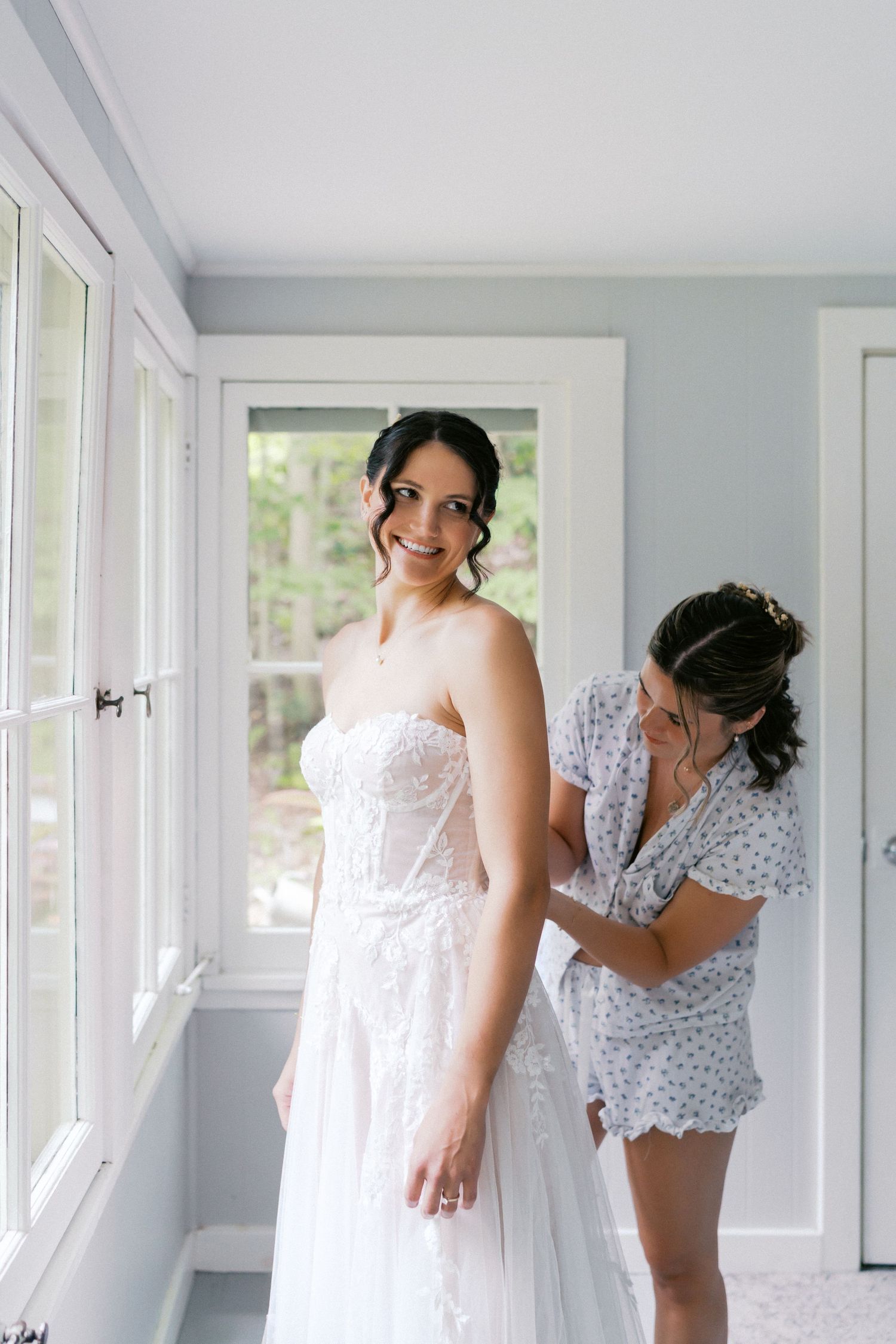 A bride getting ready in a sunlit room wearing a white lace wedding gown while someone helps with final adjustments.