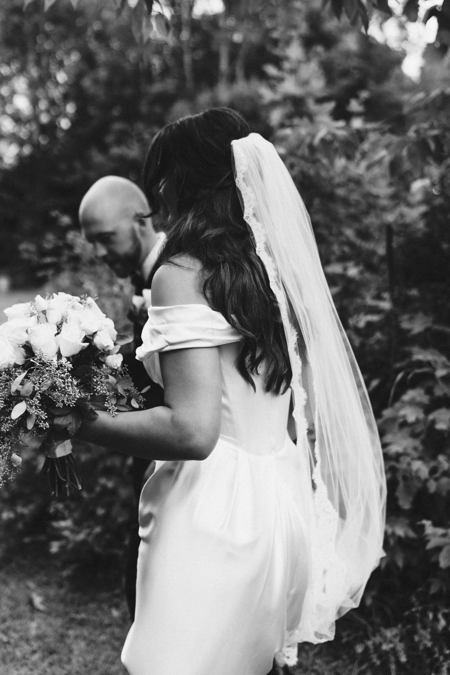 Black and white photo of a bride holding a bouquet in an off-shoulder dress with flowing veil.