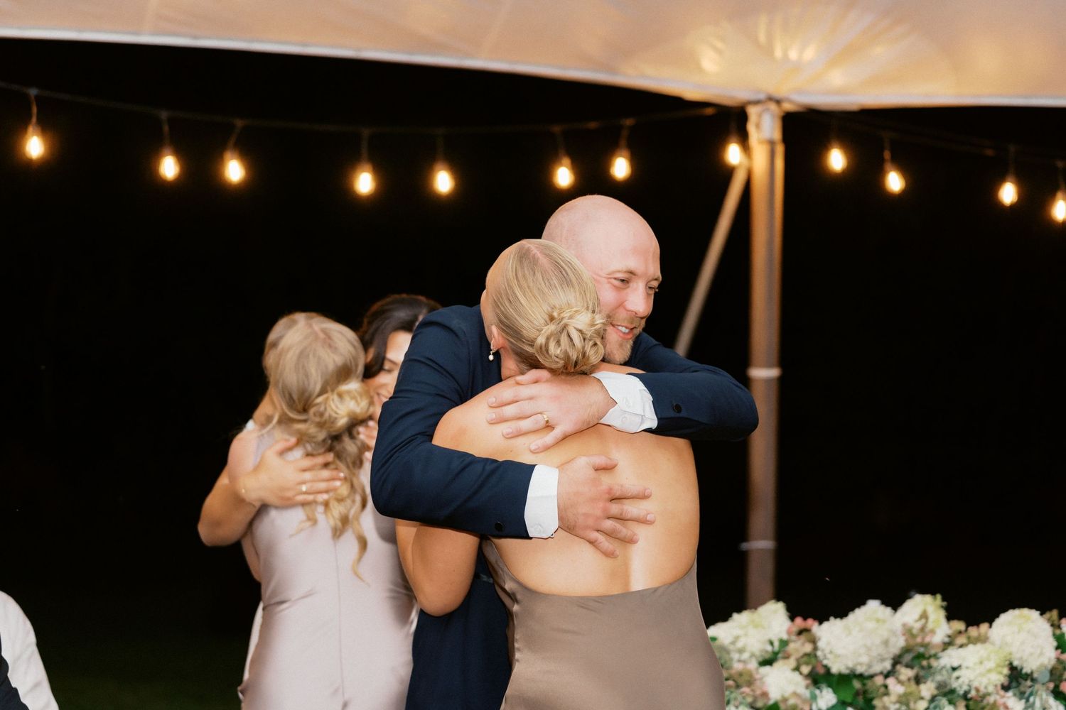Wedding guests dance and celebrate under warm string lights at an evening reception.