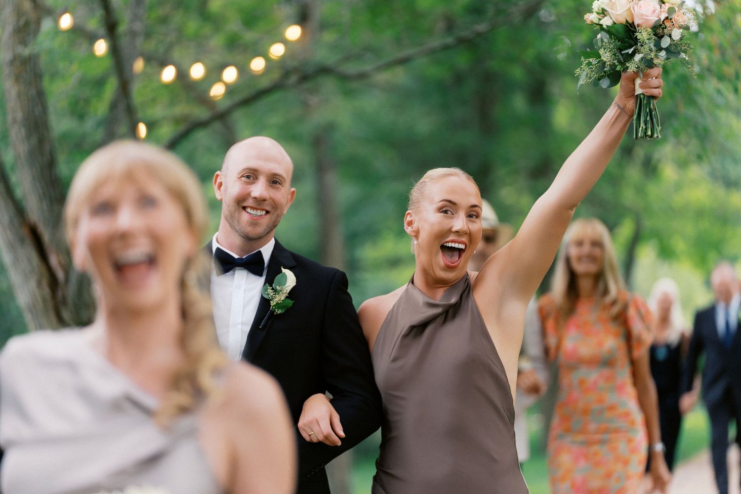 Wedding party celebrates joyfully outdoors with raised arms and laughter during a summer garden reception.