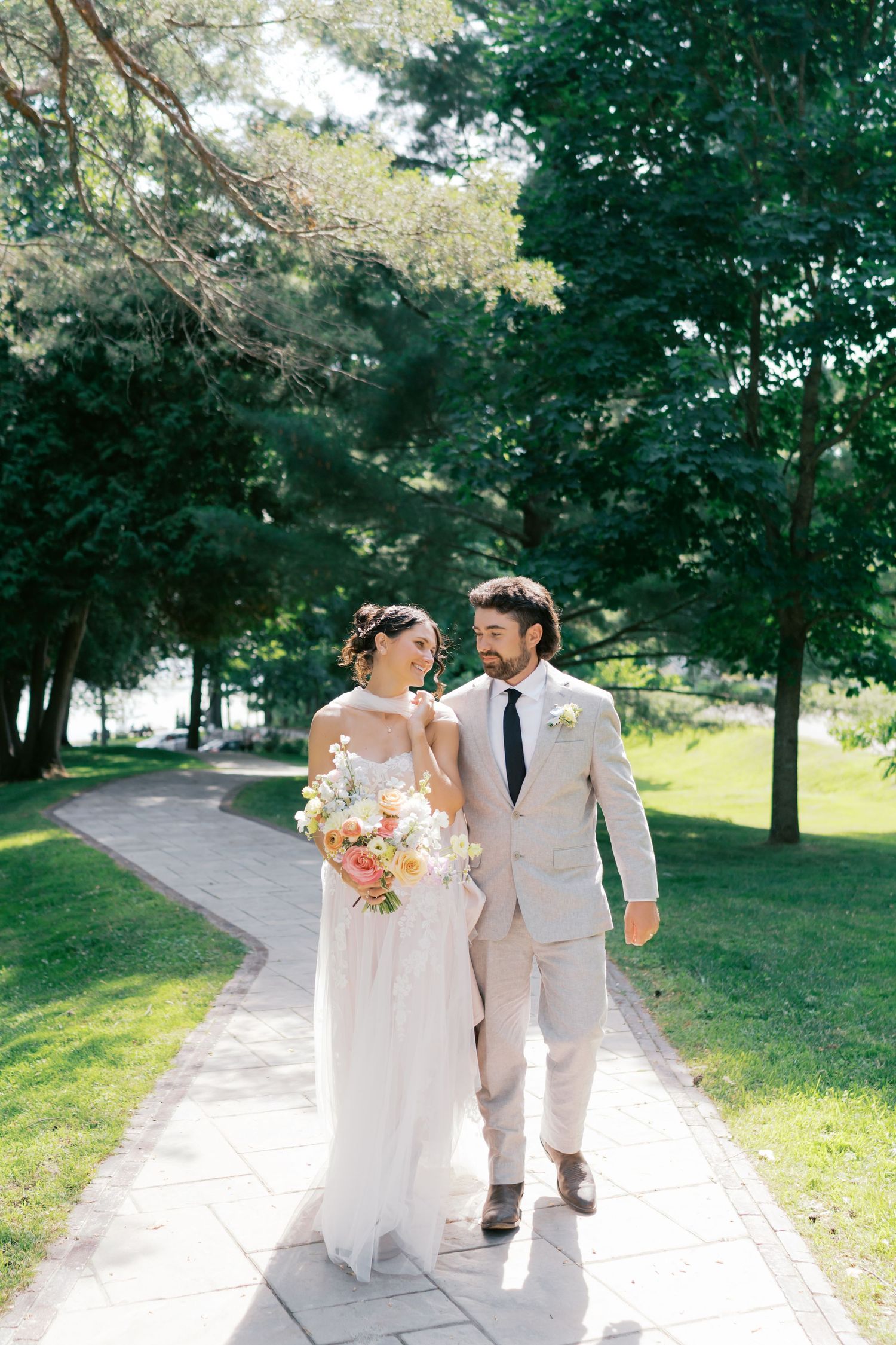 Newlyweds walking together on a tree-lined path in a sunny park.