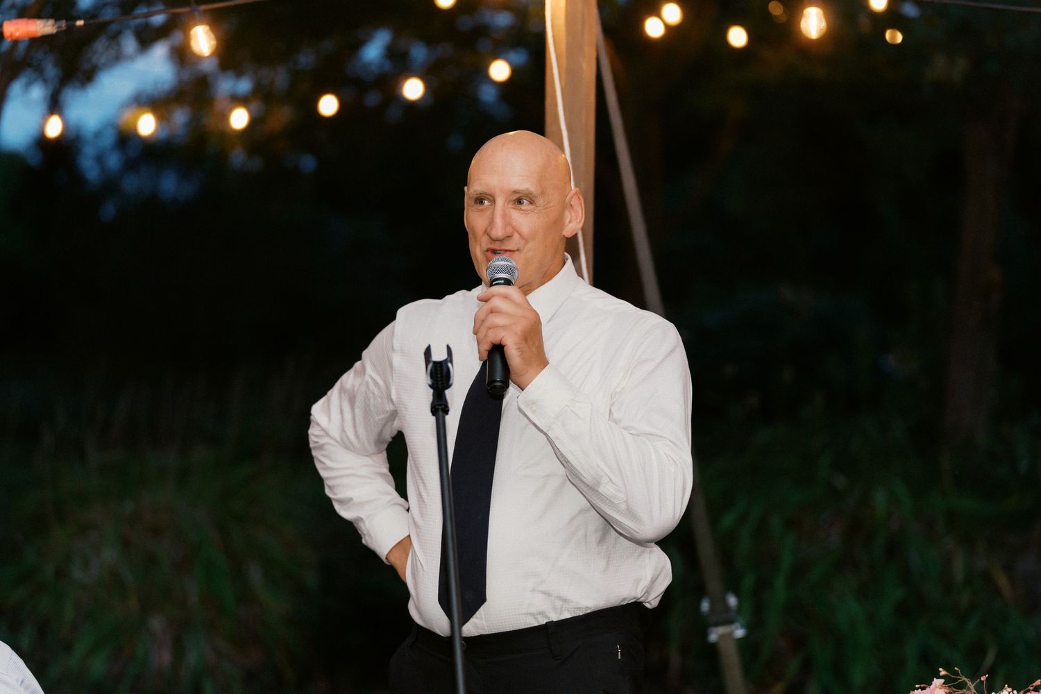 Evening outdoor reception with string lights as speaker addresses wedding guests.