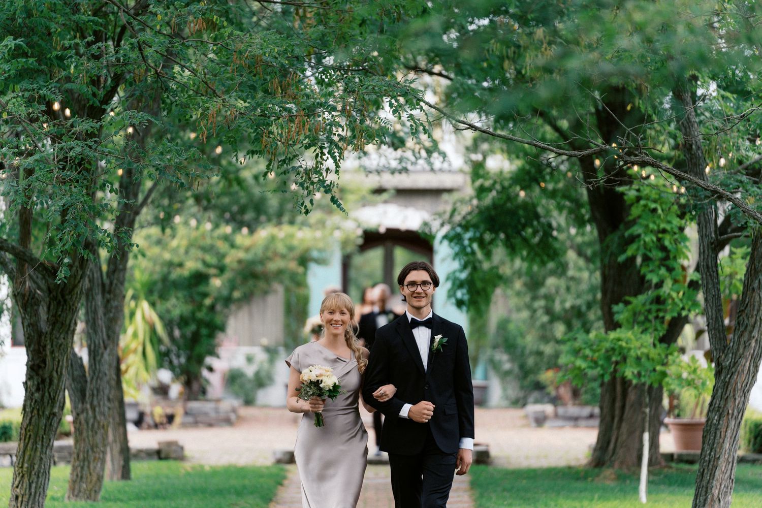 Wedding party walking through a tree-lined path in formal attire during an outdoor ceremony.