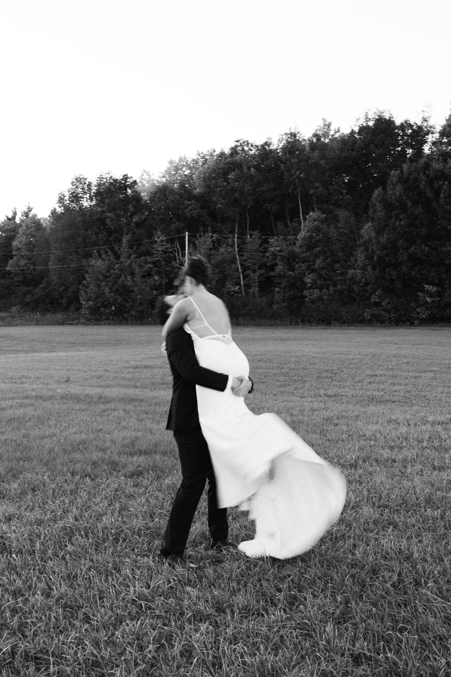 A bride and groom dance and twirl in a grassy field with trees in the background.
