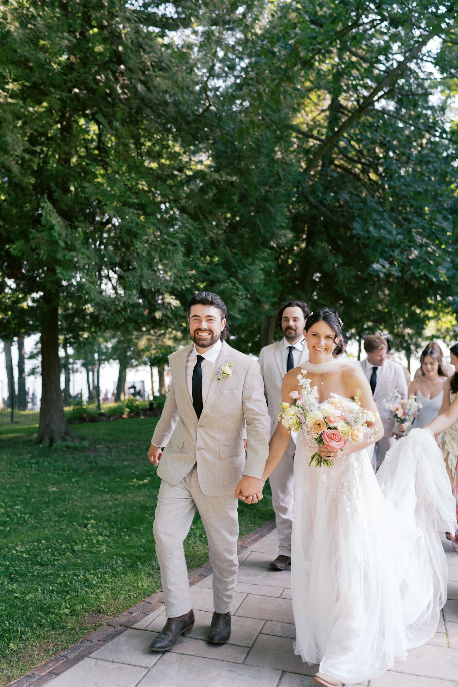 Wedding party in light colored attire walking together outdoors on a tree-lined path.
