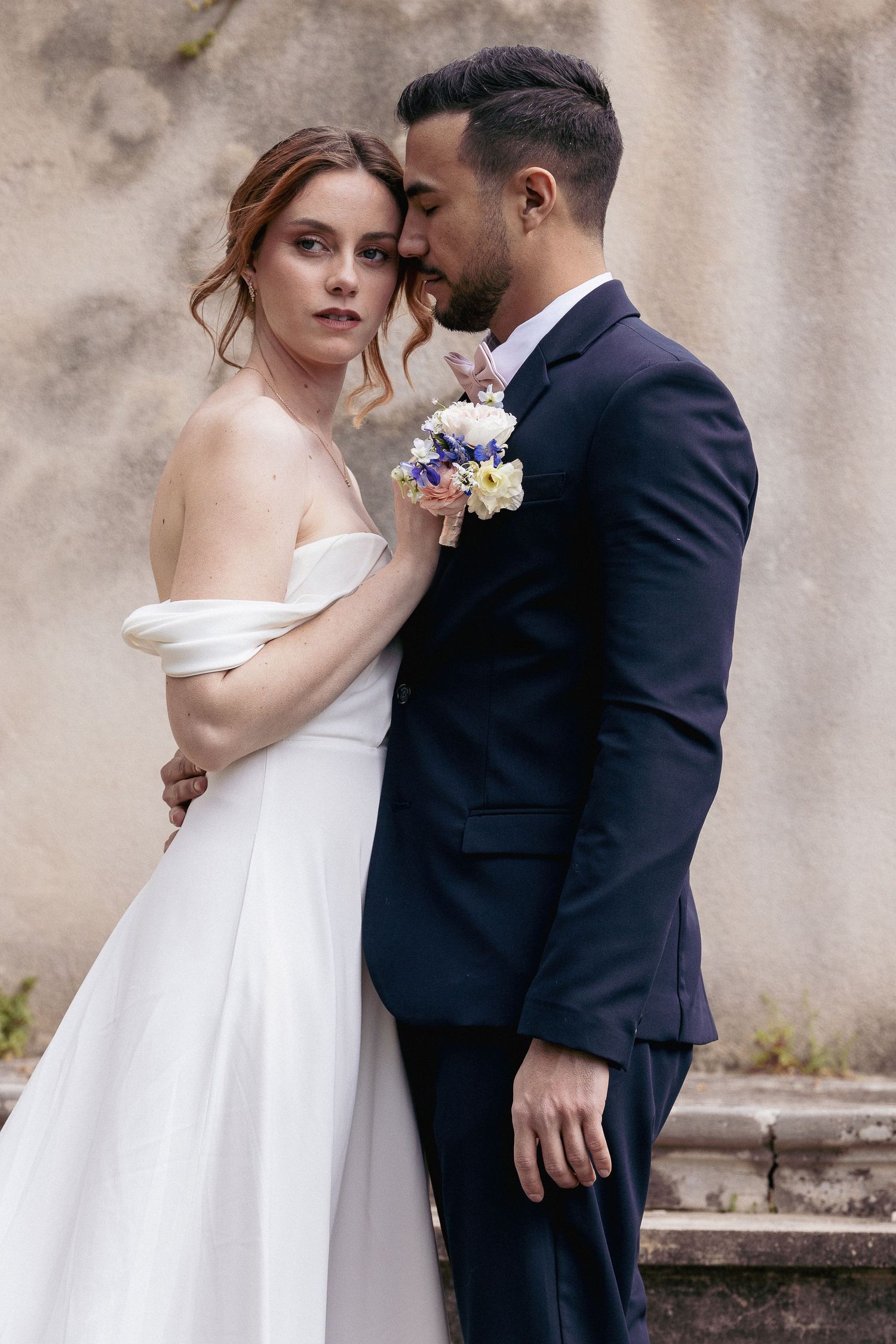 Elegant bride in off-shoulder gown and groom in navy suit pose against textured wall.