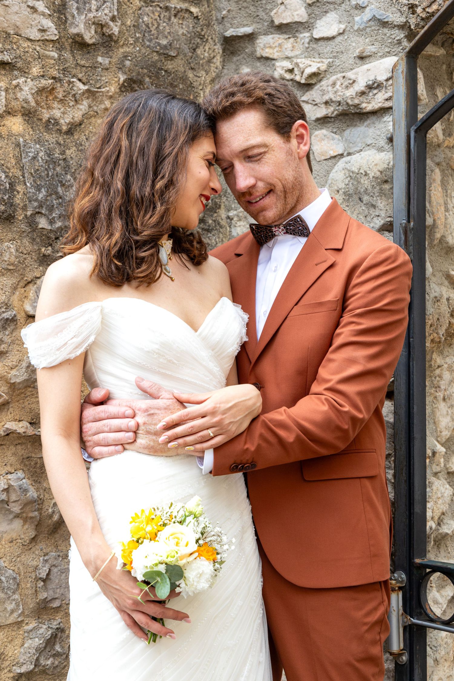 Romantic wedding couple embraces against stone wall as bride holds yellow and white bouquet.
