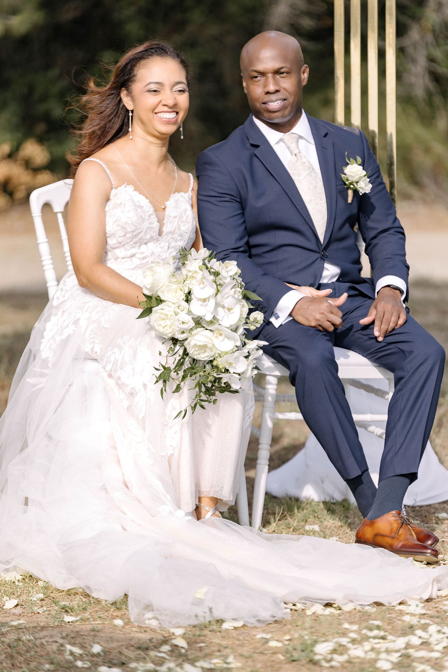 Elegantly dressed couple poses on white chairs at their outdoor wedding.