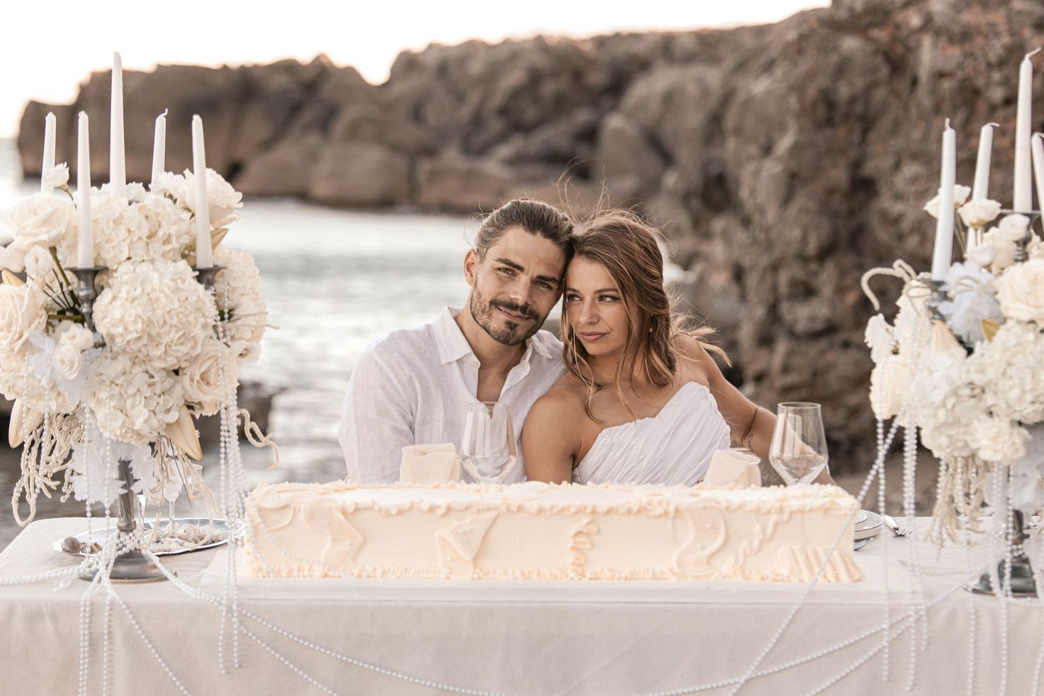 Couple poses at elegant white floral beach wedding table with candles and cake by rocky ocean backdrop.