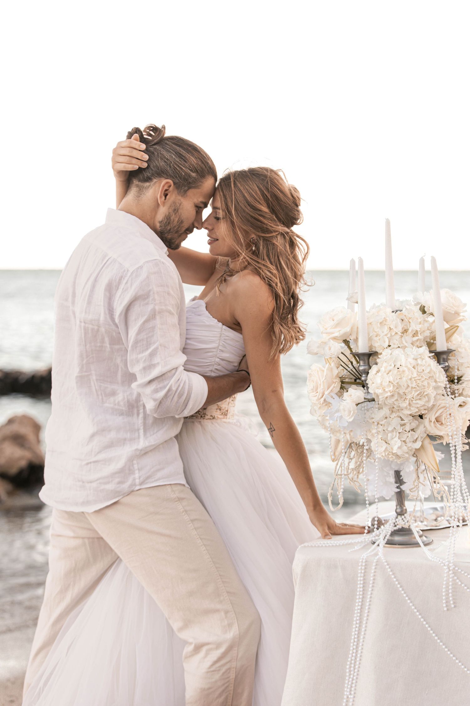 Couple in white attire sharing a romantic moment by the ocean at a beach wedding.