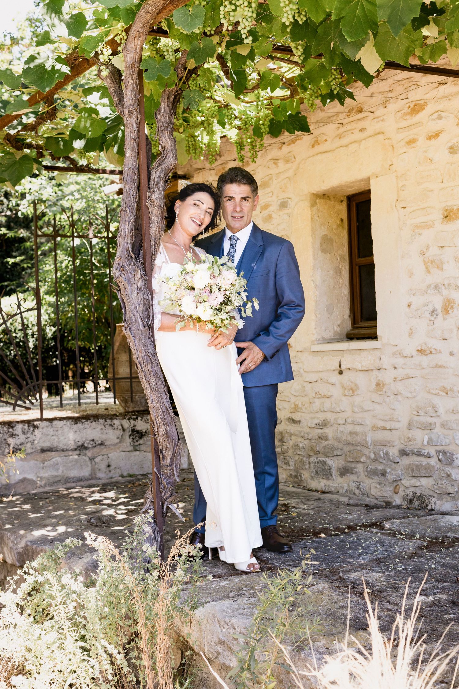 Wedding couple poses under green foliage near a rustic stone building.