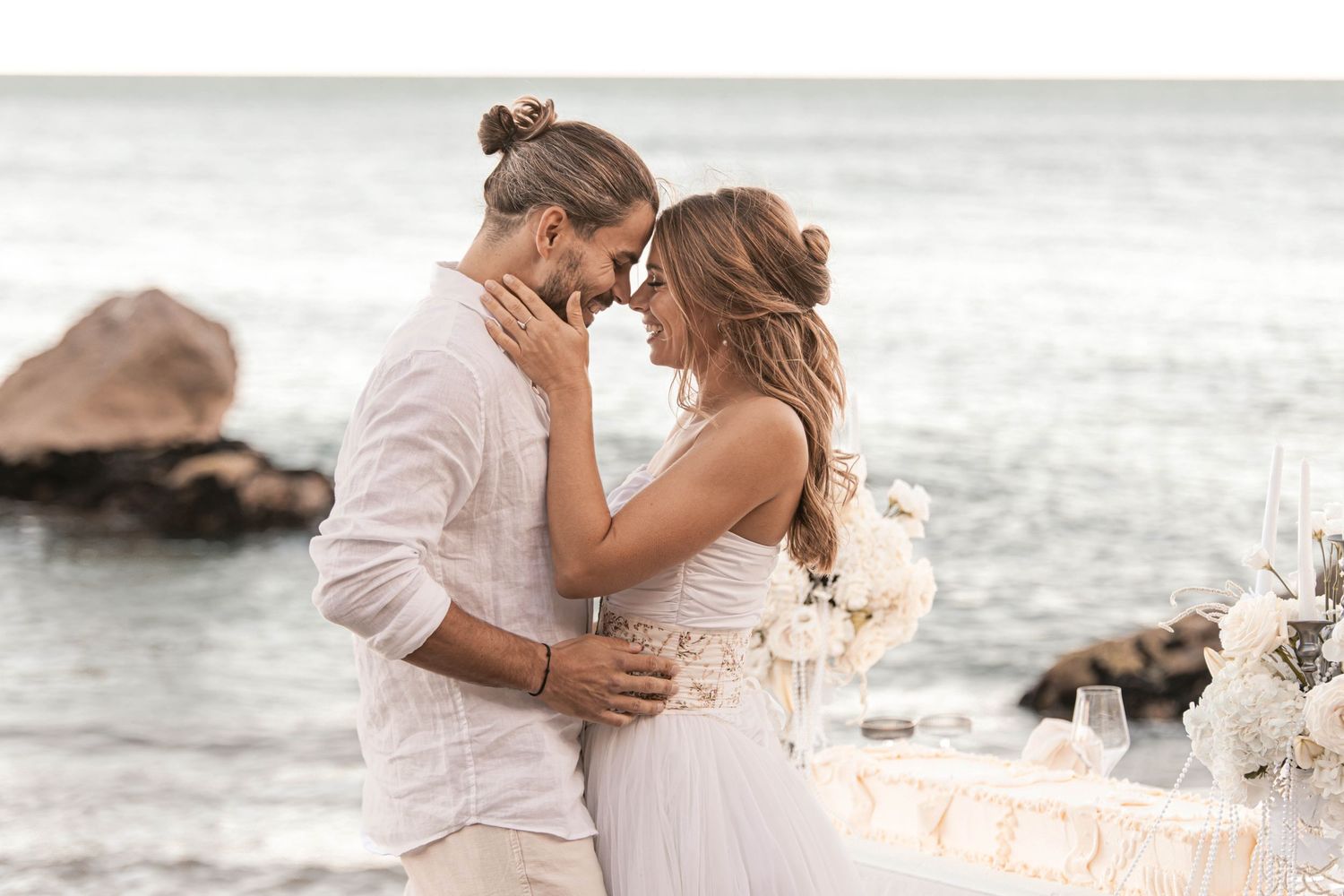 Romantic couple embracing at beach wedding ceremony with ocean backdrop, bride in white dress and groom in linen suit.