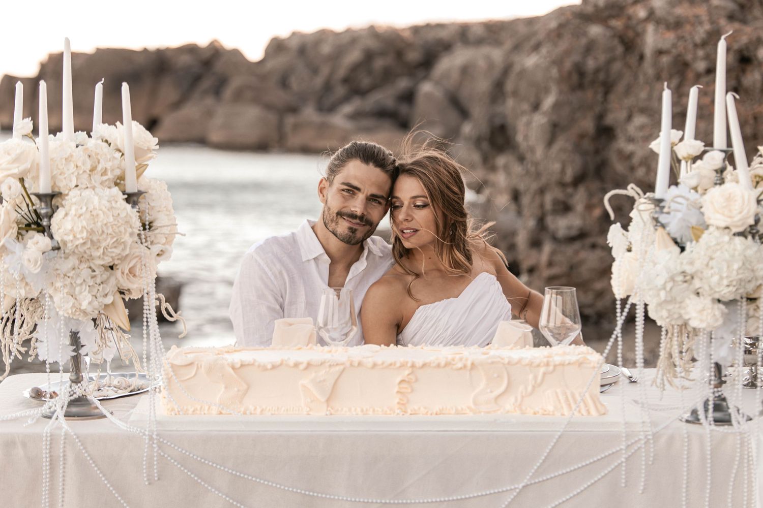 Couple sits at elegant beachside wedding table with white flowers, candles, and a tiered cake by rocky ocean backdrop.