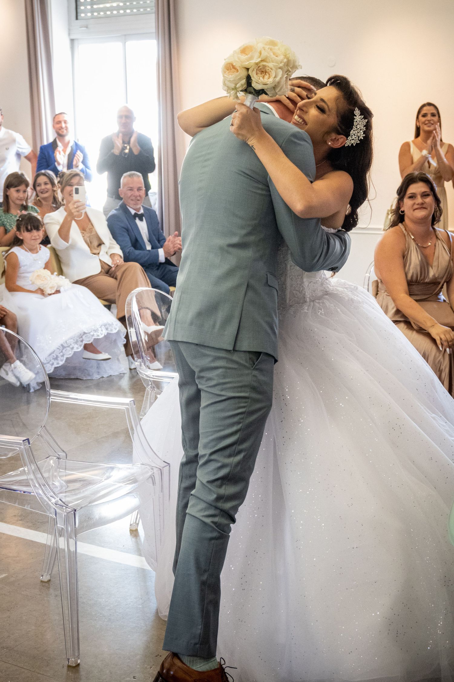 A joyful wedding couple embraces during a dance while guests look on and applaud.