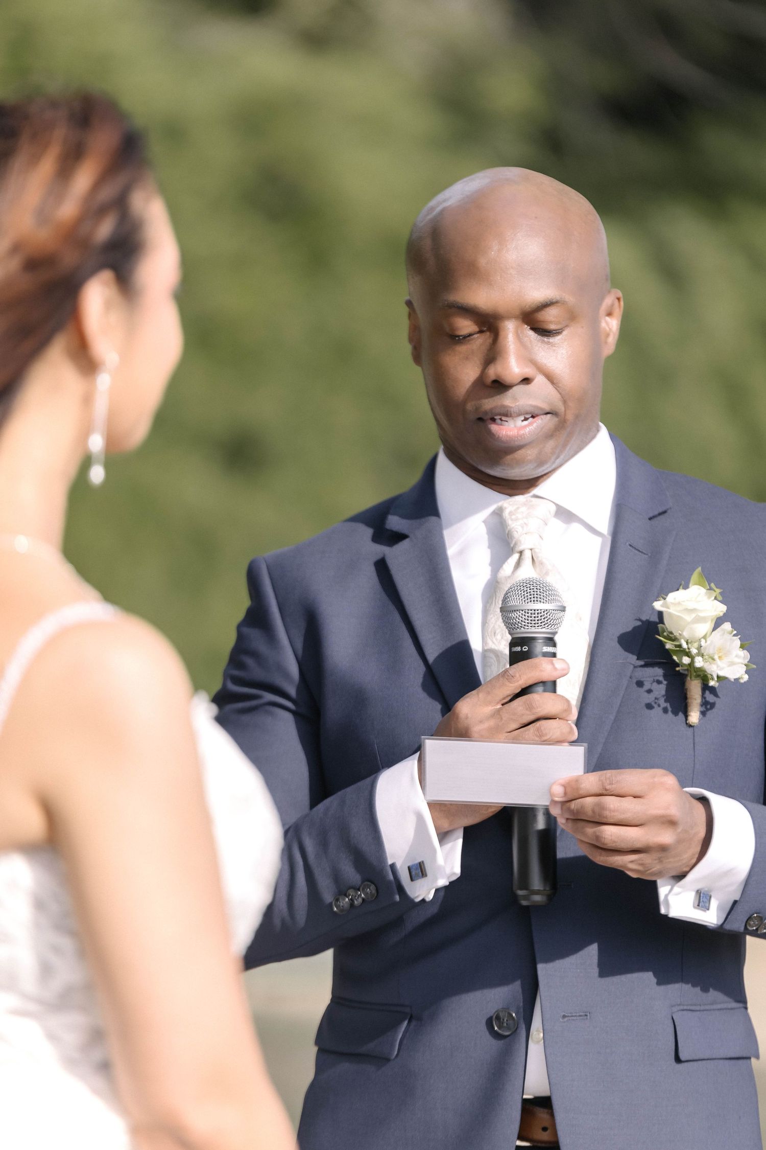 Groom reads vows from paper while holding microphone during outdoor ceremony.