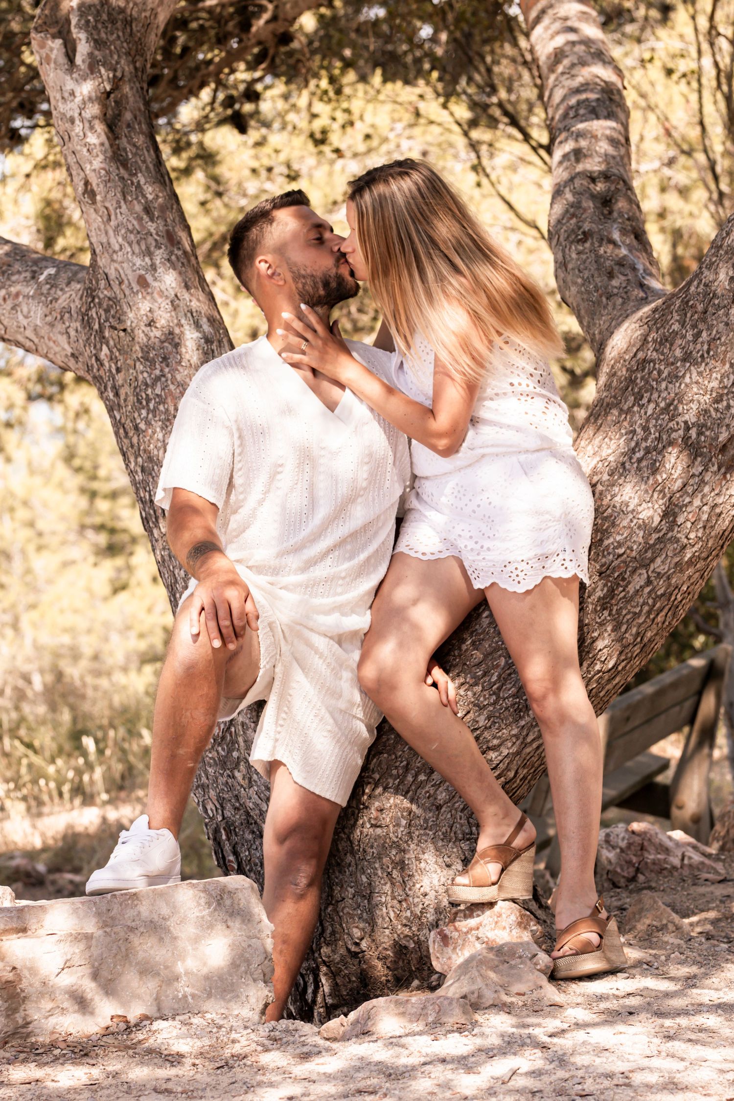 A couple in white outfits shares an intimate moment against a tree trunk in a romantic outdoor photoshoot.