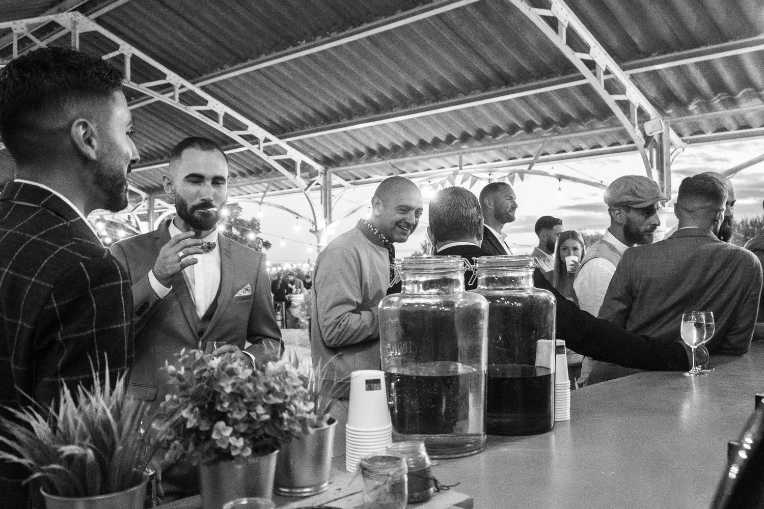 Black and white photo of guests gathering around drink dispensers at outdoor reception.
