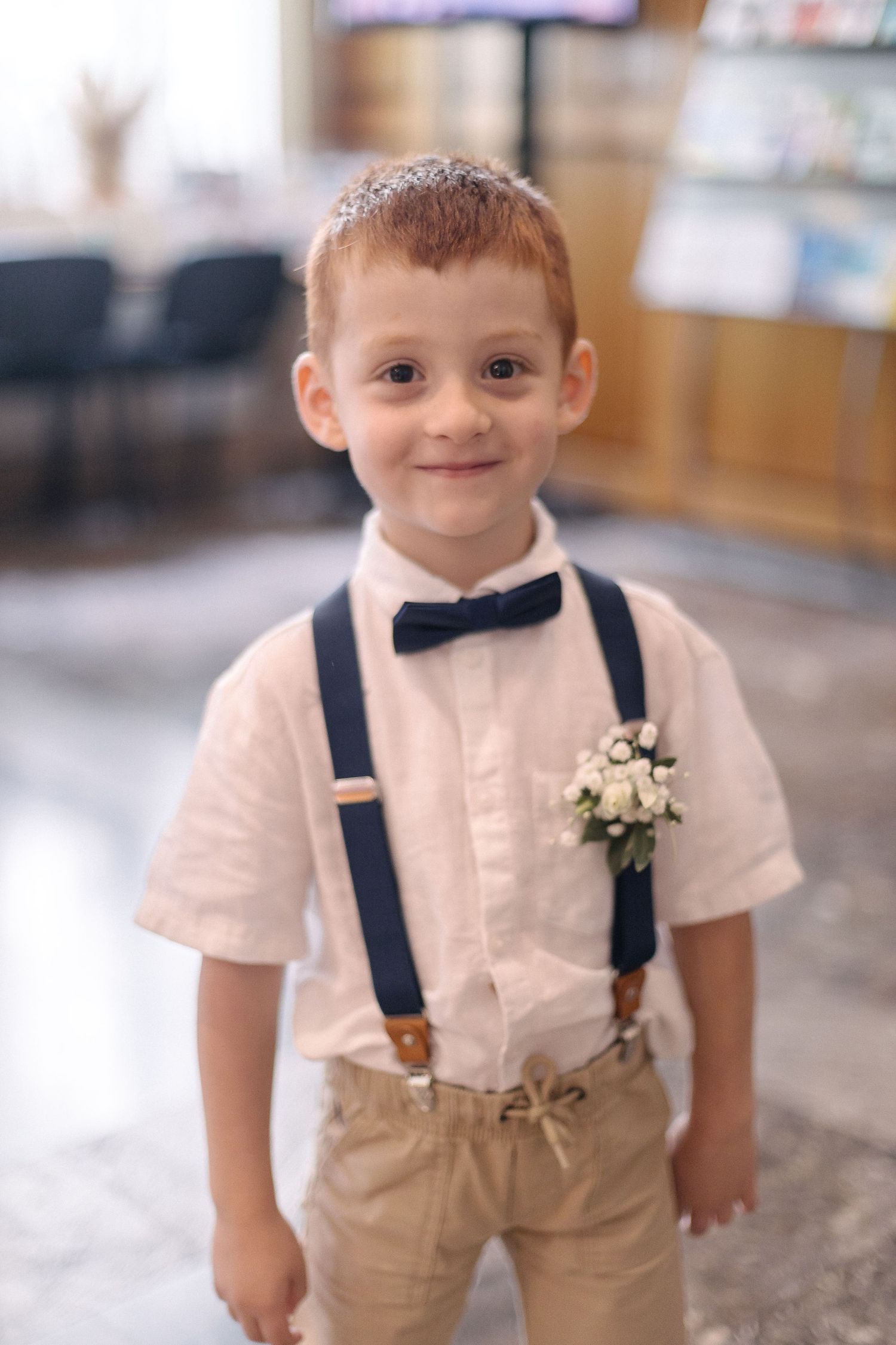Adorable ring bearer in white shirt, suspenders and bow tie smiles at wedding ceremony.