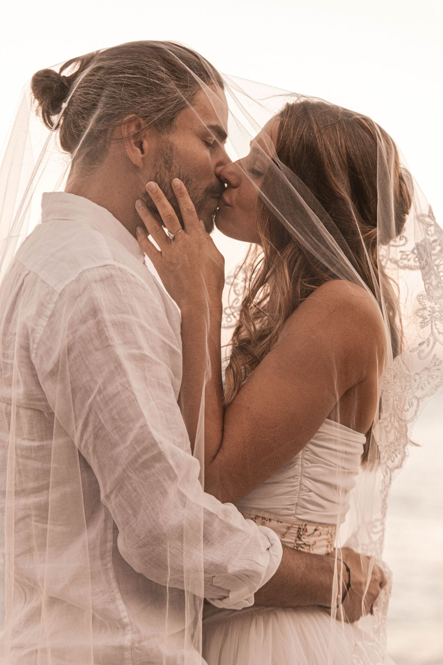 A bride and groom share a romantic kiss under a lace wedding veil on a beach.