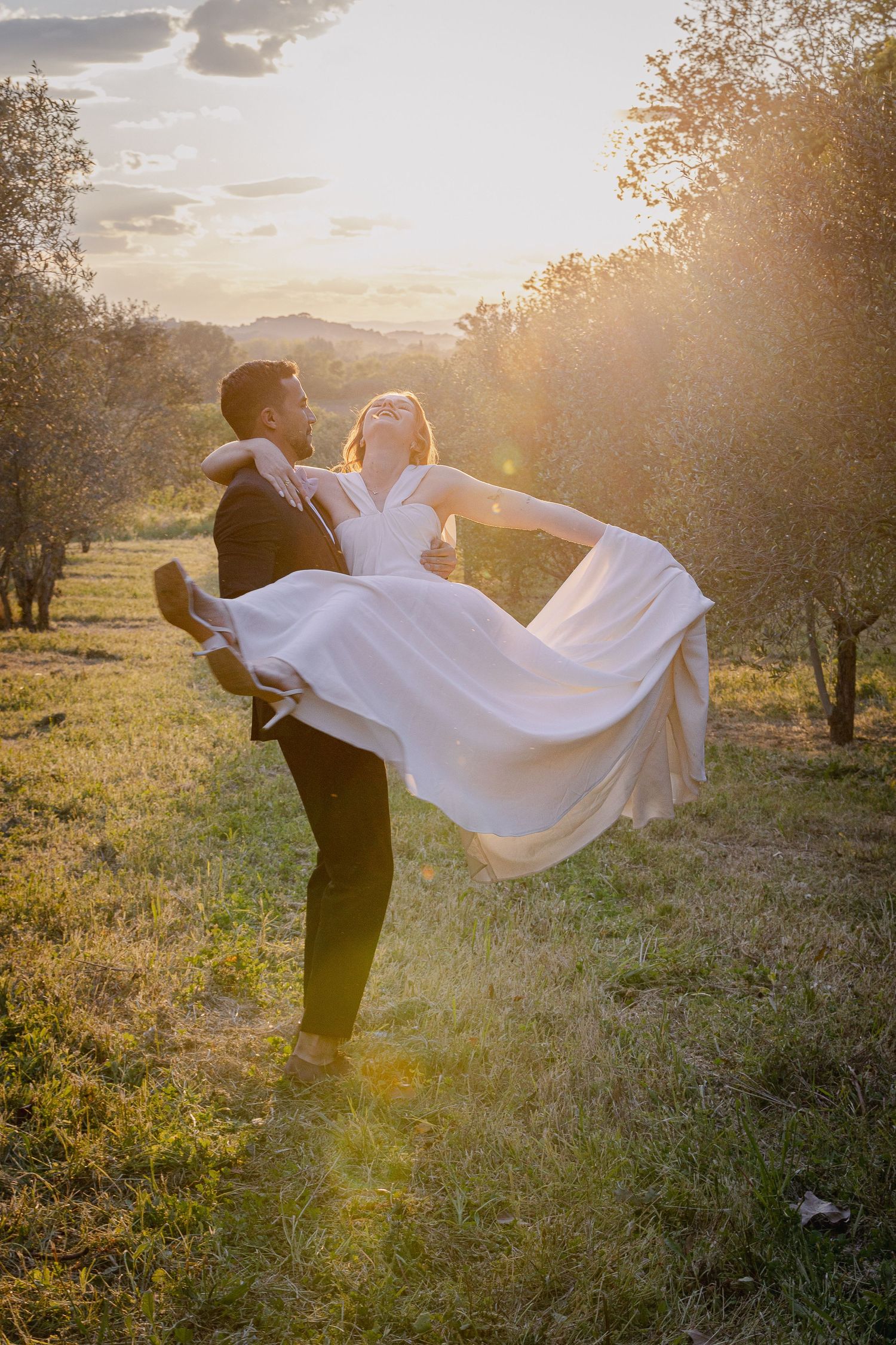 Groom lifts bride in flowing white dress during sunset in rustic outdoor setting.