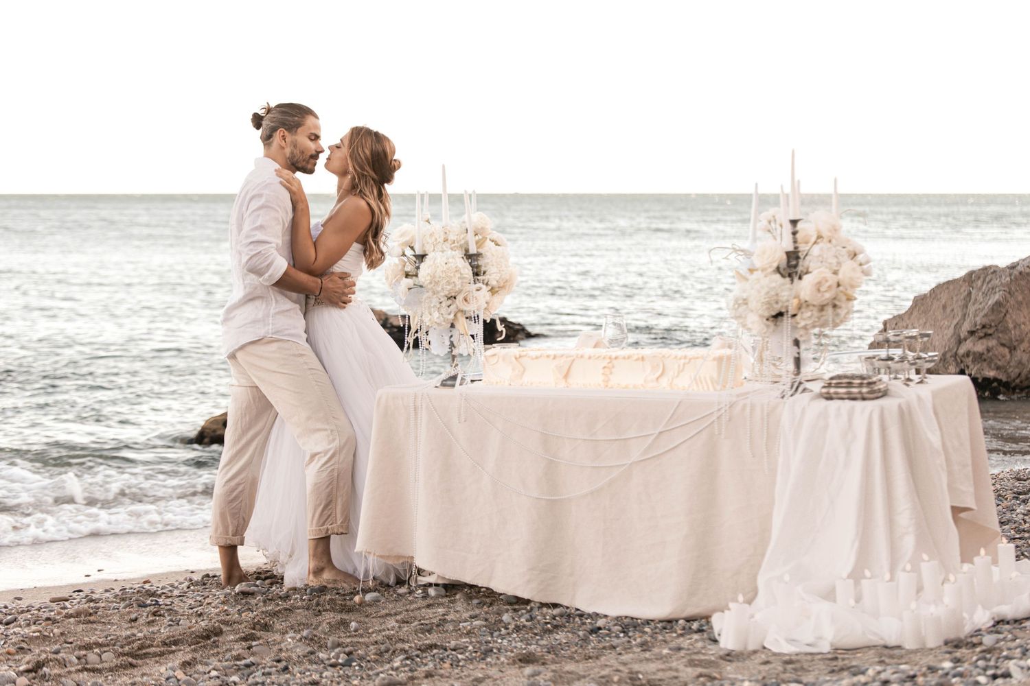 Couple in white attire embracing at a romantic beach wedding setup with floral arrangements and candles.